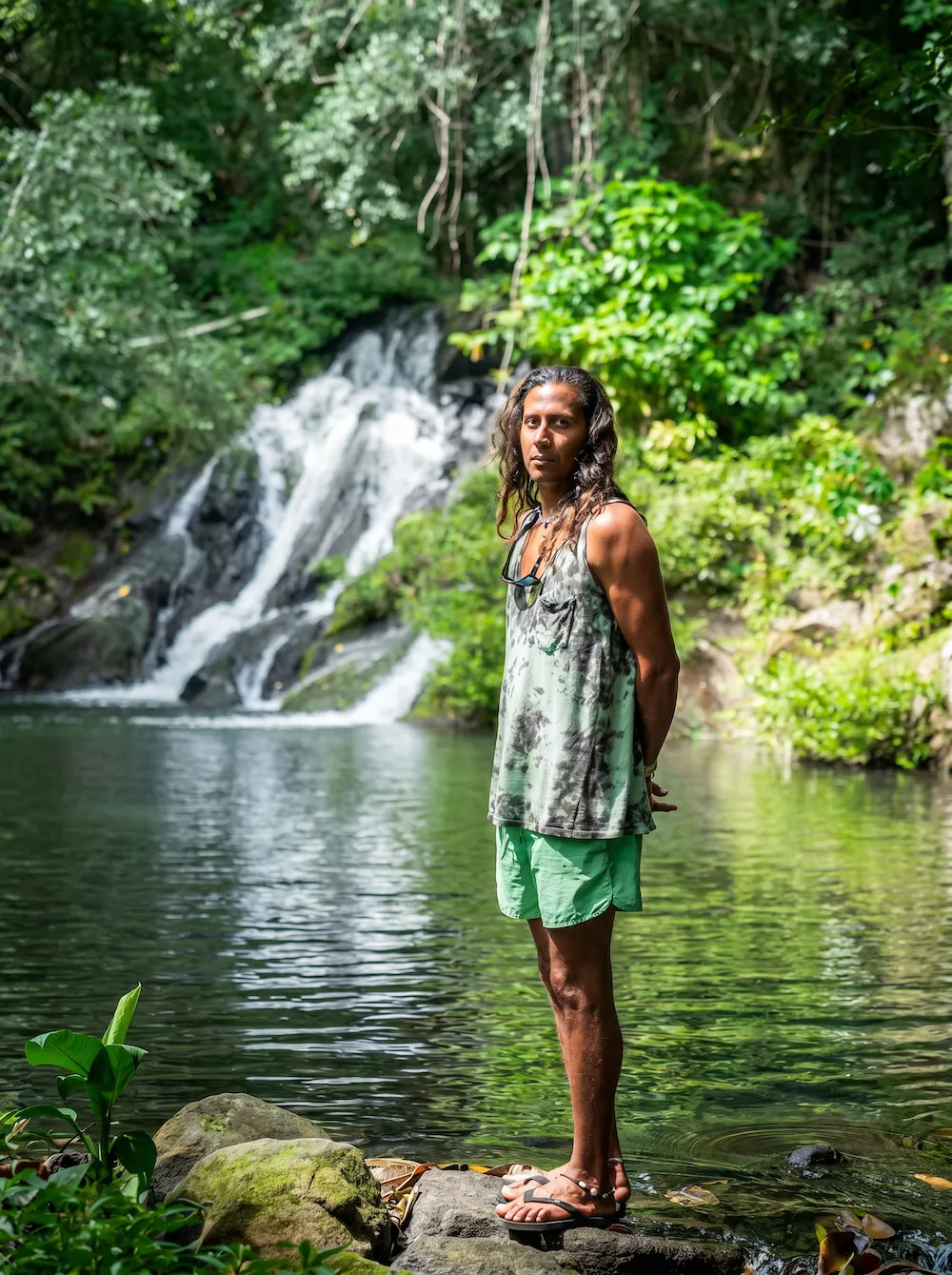 Person with long hair in green shorts standing on rocks by a forested waterfall pool.