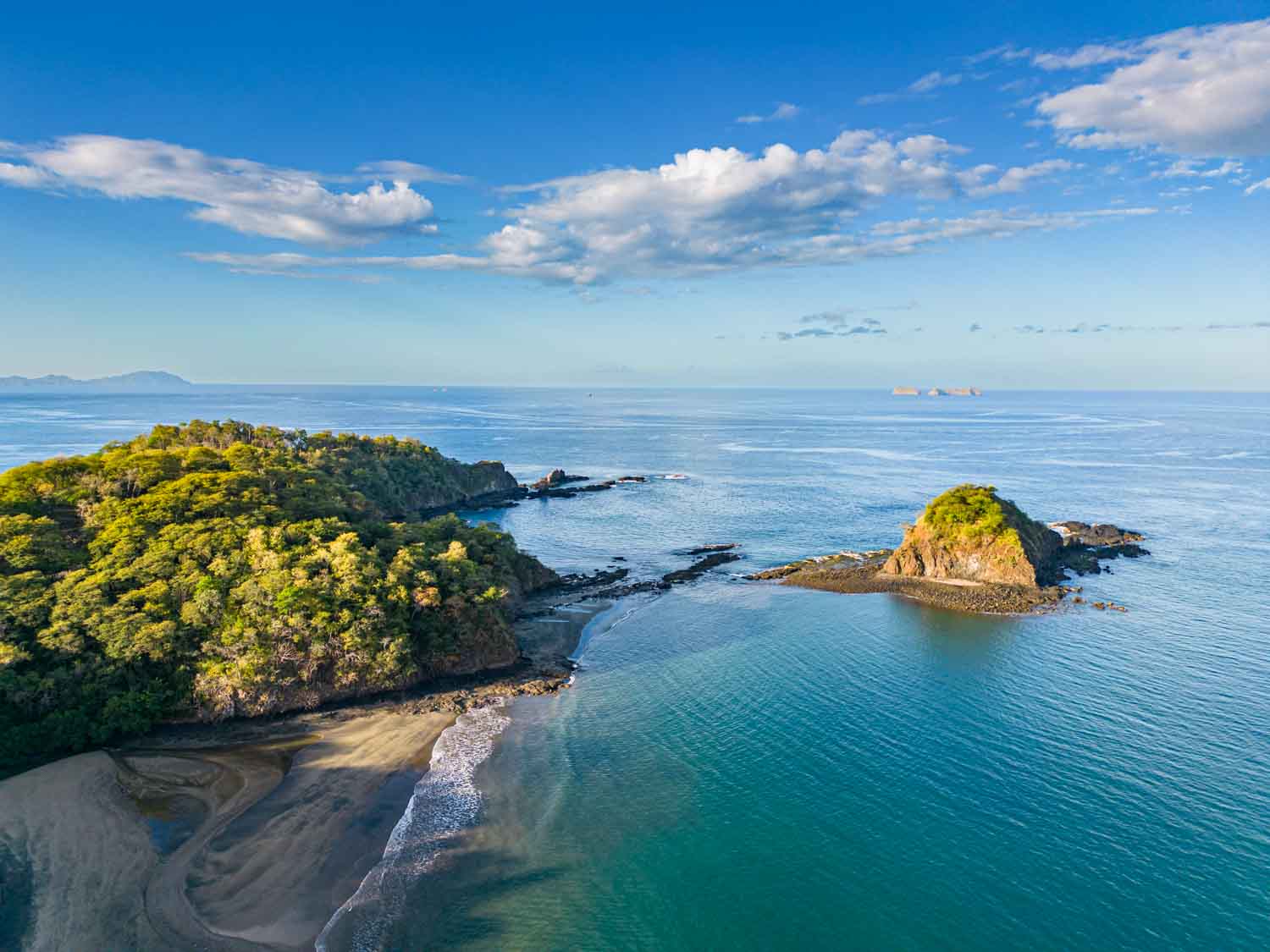 Coastal scene with two forested rocky islands and a sandy beach under blue sky with scattered clouds.