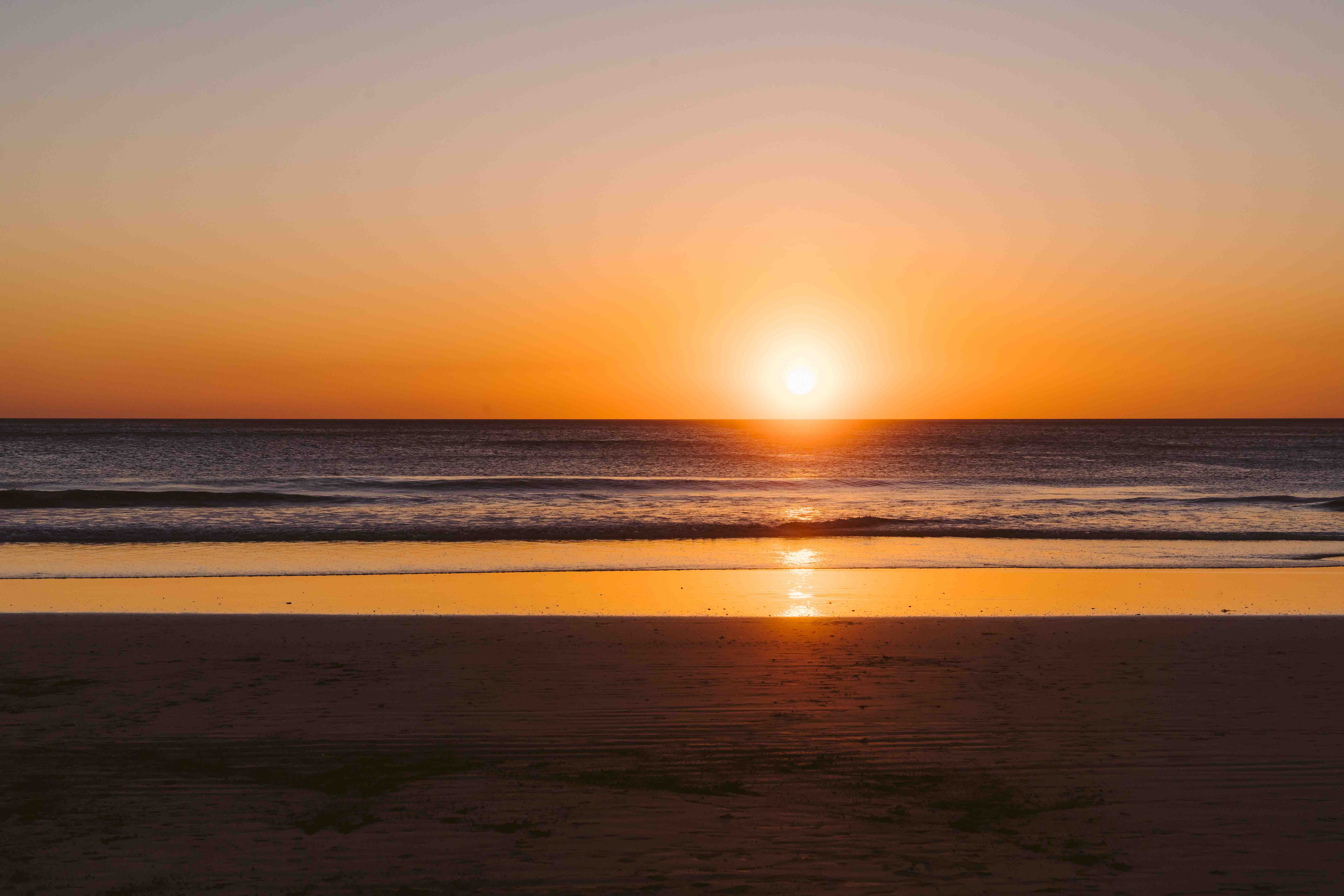 Sun setting over the ocean with waves and a sandy beach in the foreground.