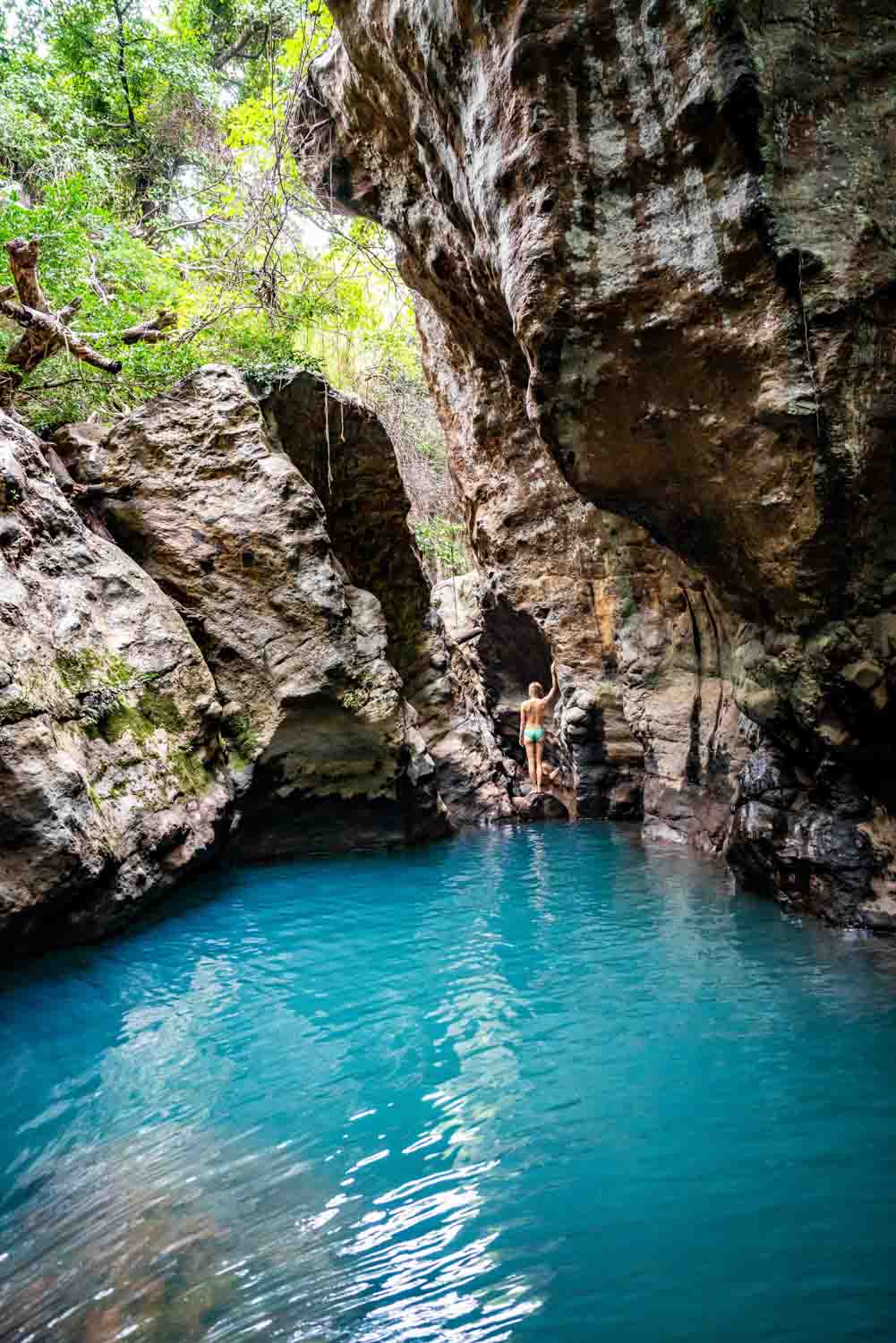 Person standing on rocks next to turquoise water surrounded by large rocky cliffs and greenery.
