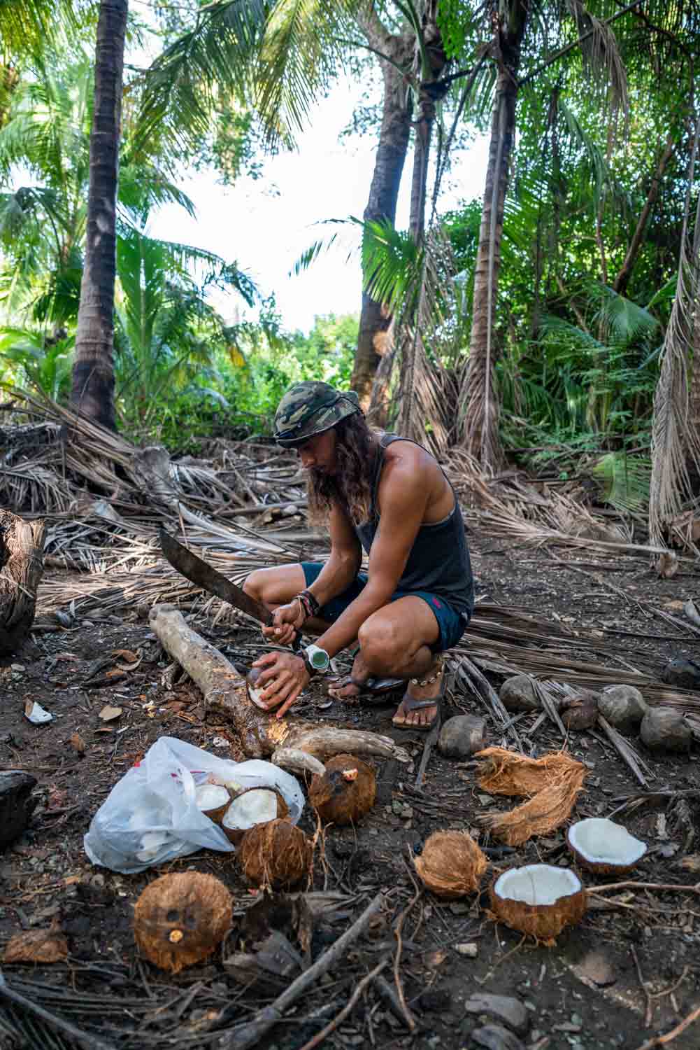 Man wearing hat and shorts using a machete to open coconuts on the forest floor.