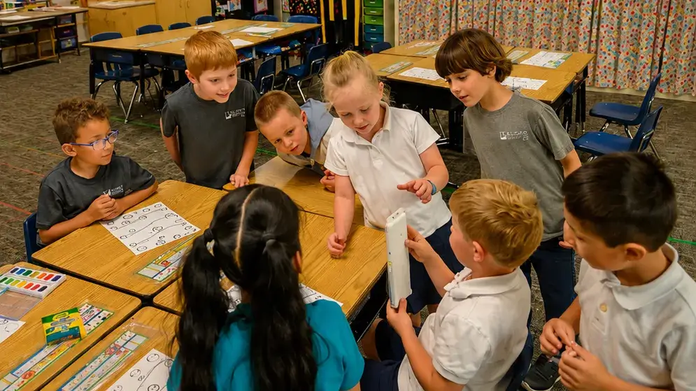 A group of young schoolchildren gathered around a boy holding a a color paint set in a classroom at St Joseph Marquette Catholic School.