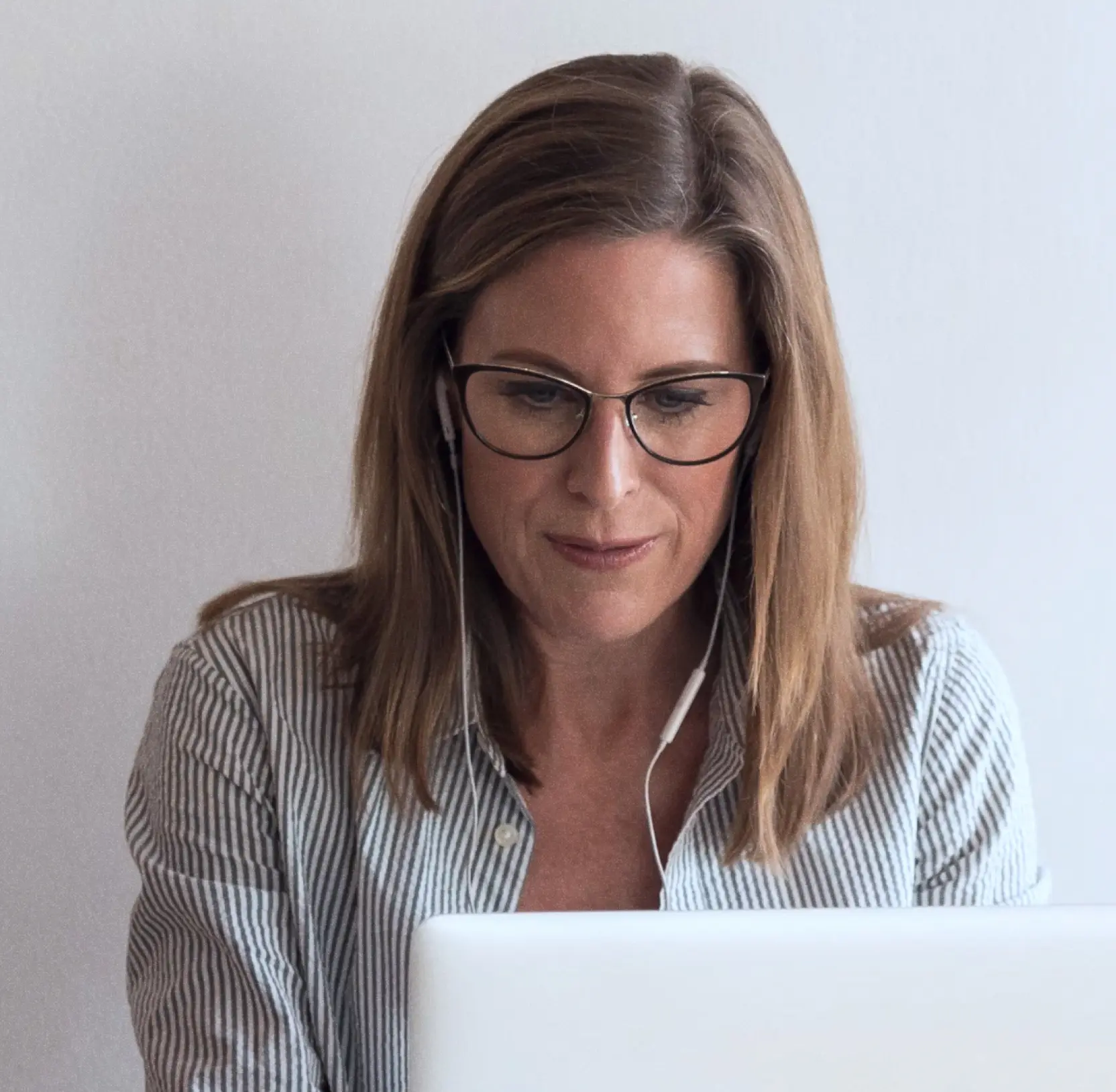 Woman with glasses wearing earbuds, looking at a white laptop against a plain background.
