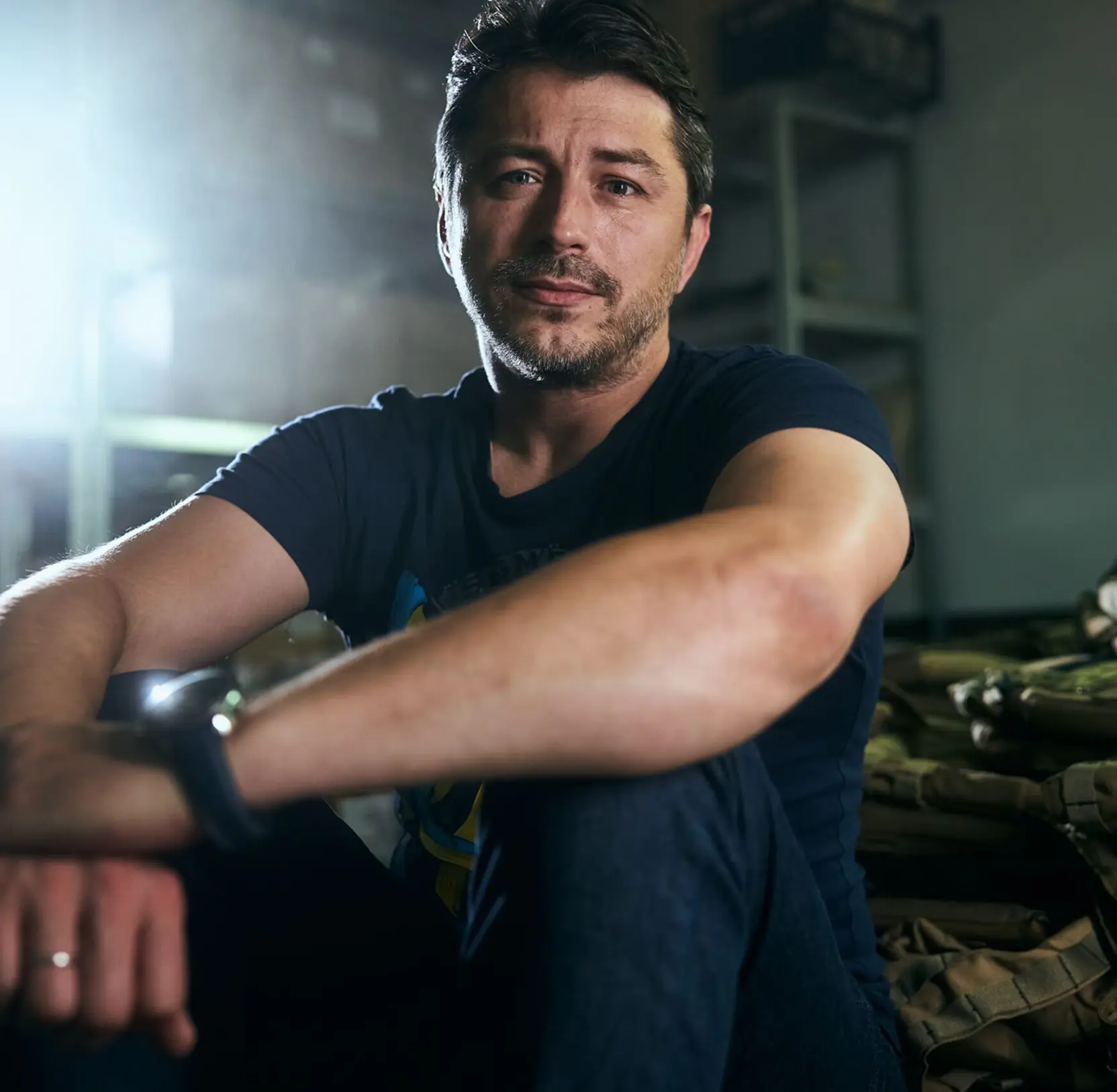 Man with short hair and beard wearing a dark t-shirt sitting with arms resting on knees in a dim room.