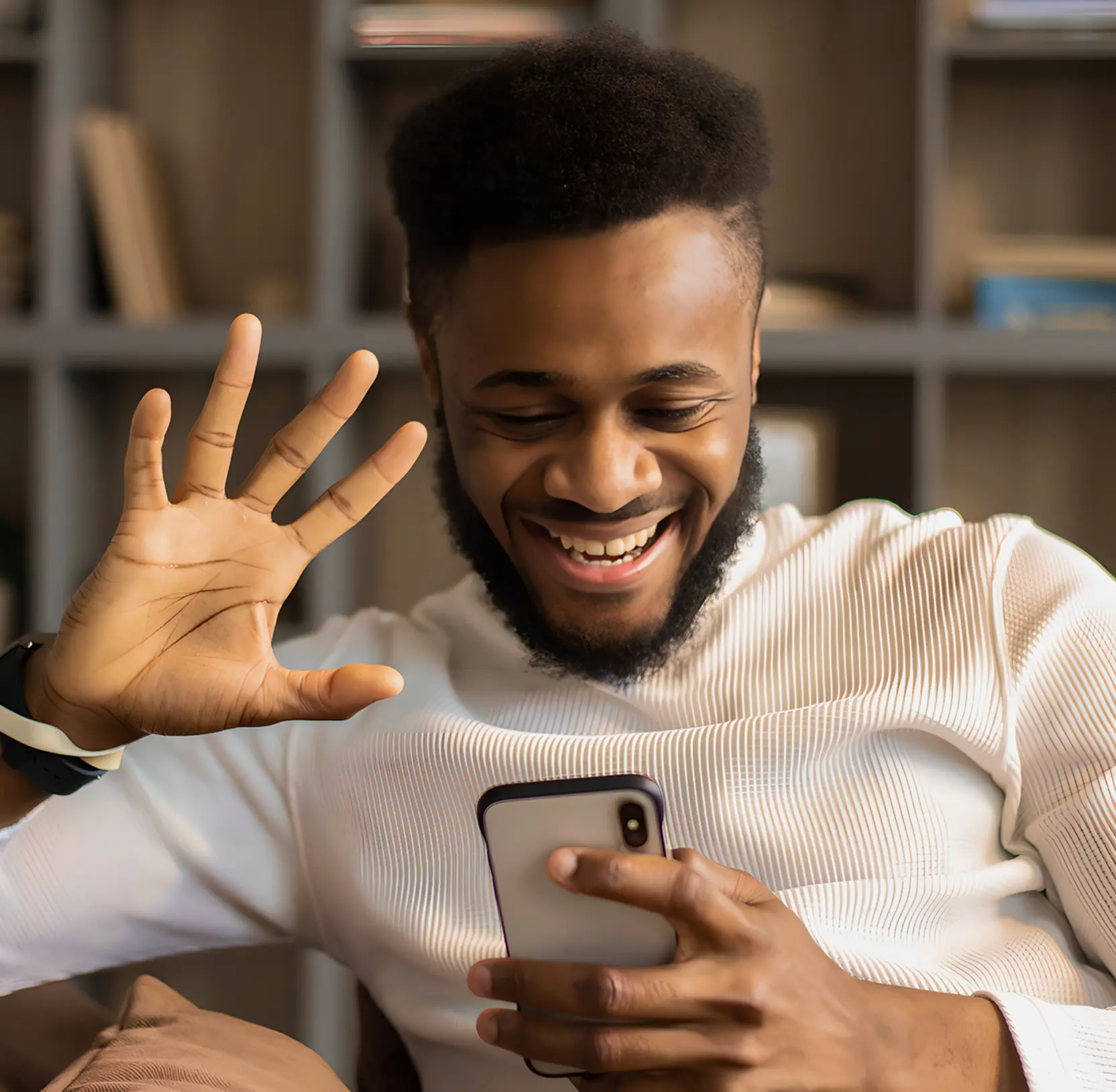 Smiling man waving with one hand while looking at a smartphone in his other hand.