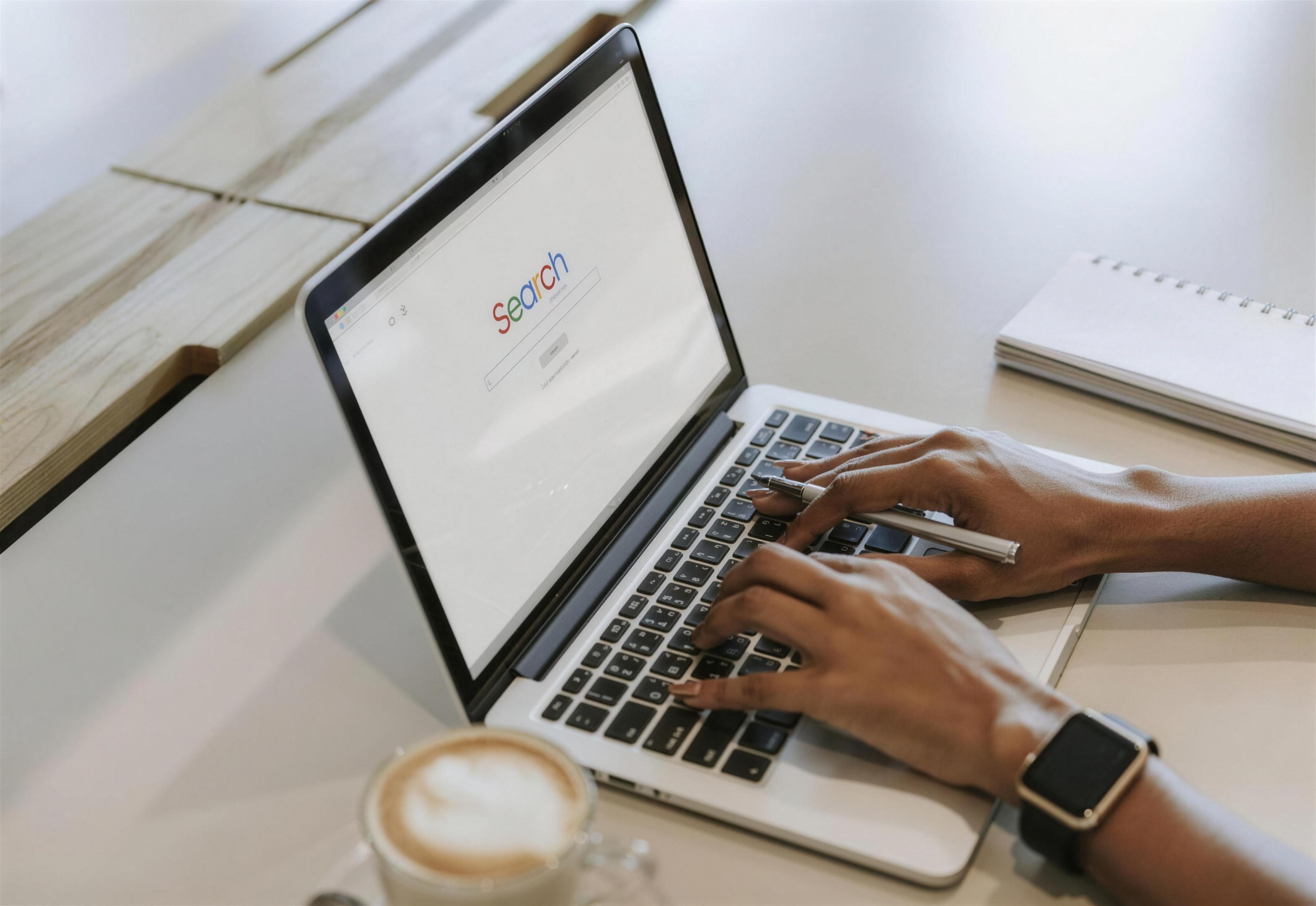 Hands typing on a laptop open to a search page, beside a notebook and a cappuccino