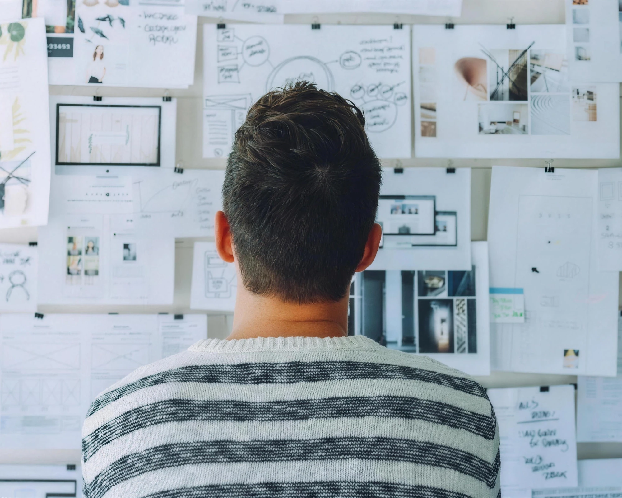 A person seen from behind studying a wall of sketches, wireframes and printed layouts.