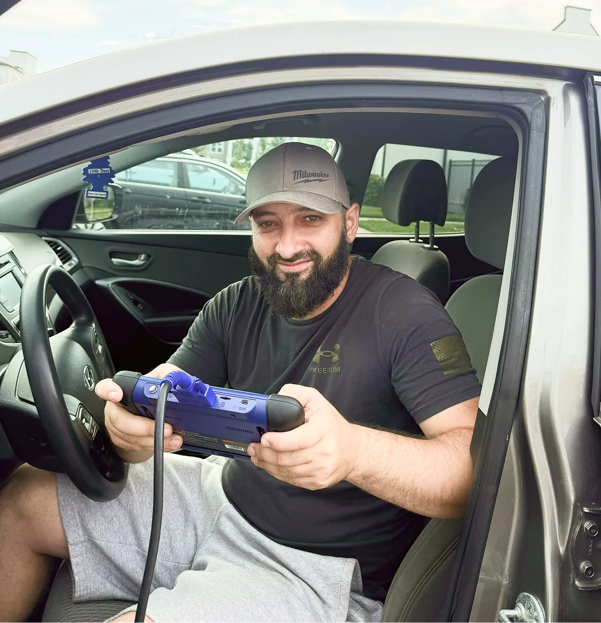 Bearded man wearing cap, sitting inside car holding a blue device with a cable attached.