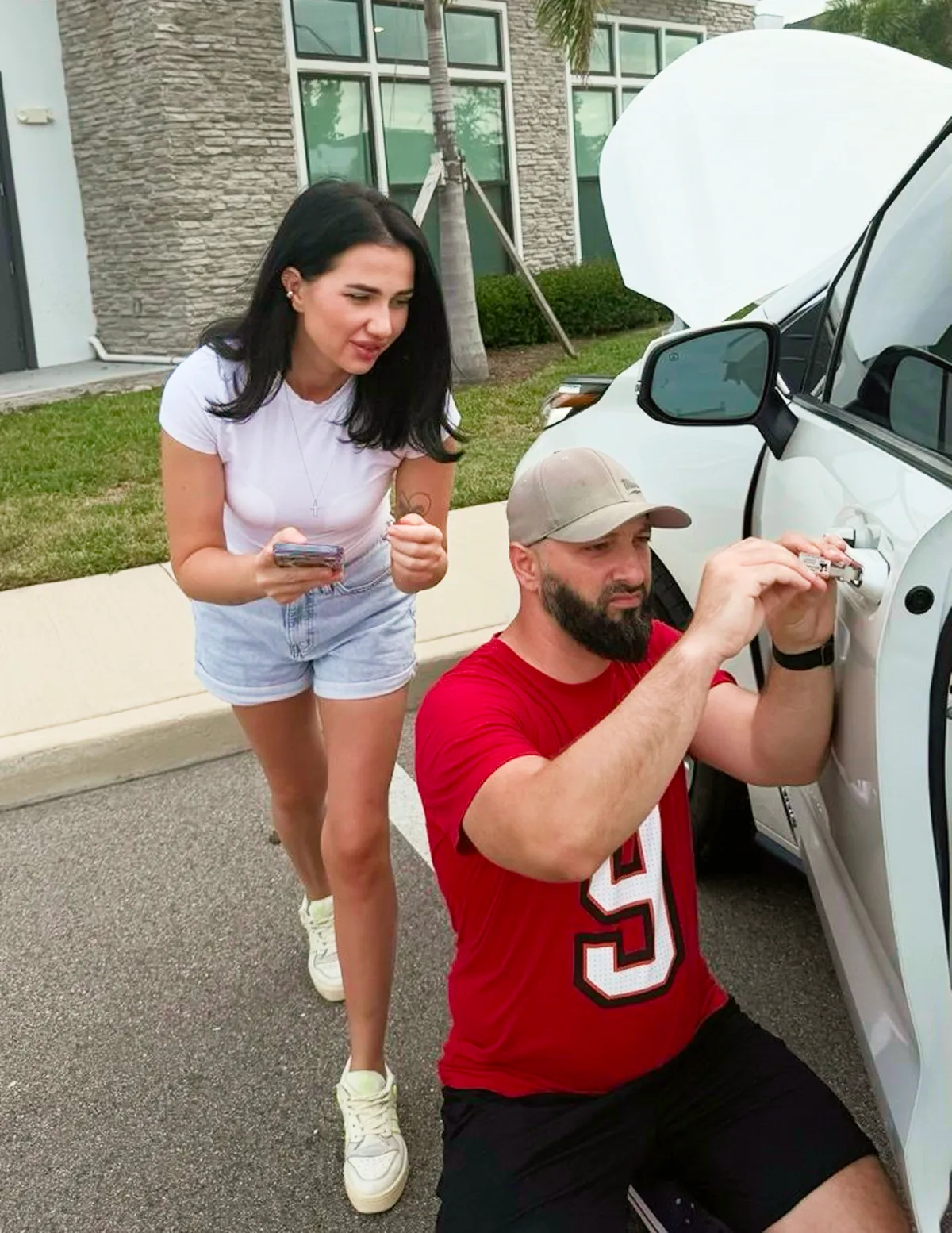 Man fixing a car door lock while a woman in shorts watches nearby outside a building.