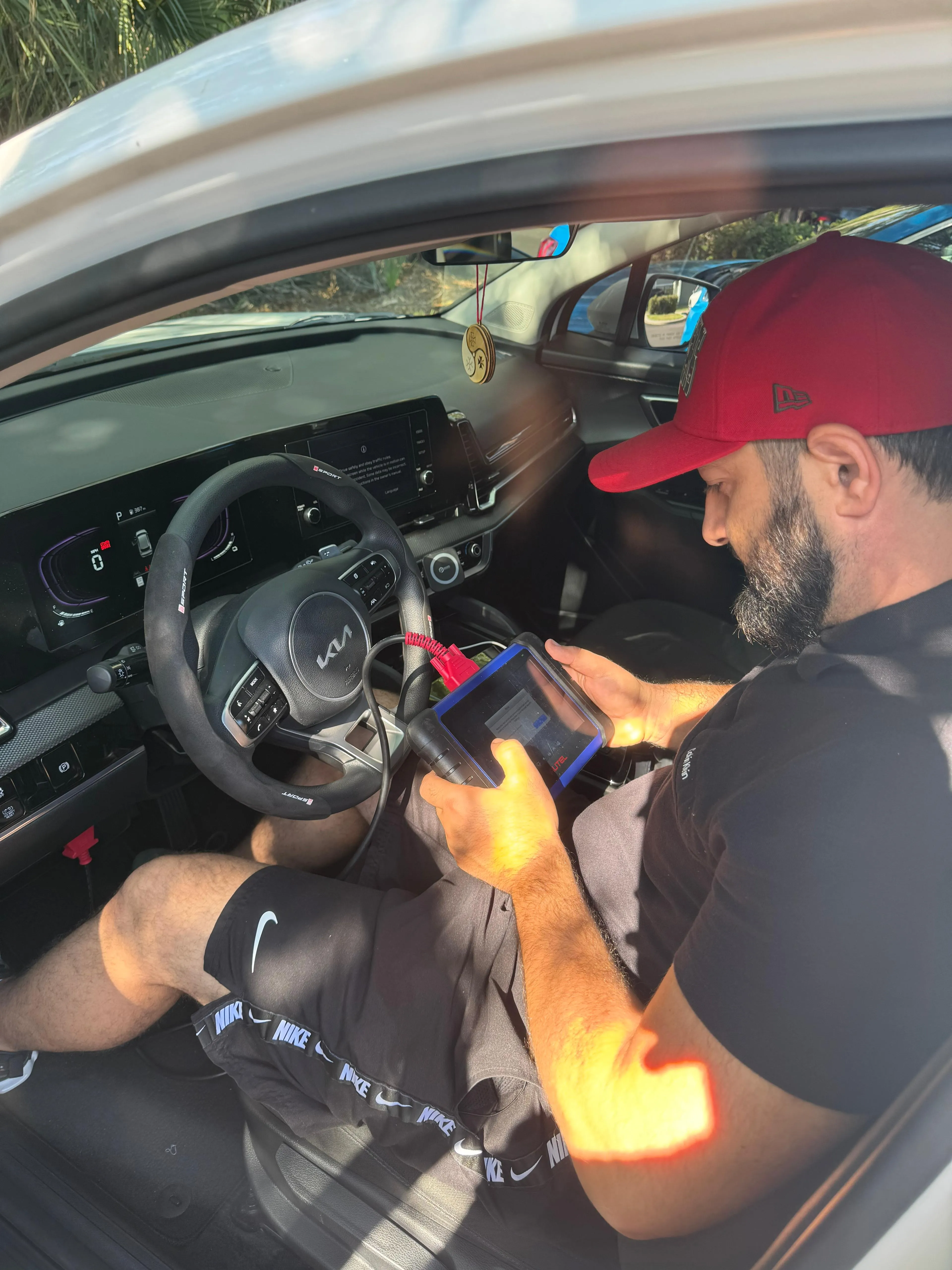 Man wearing a red cap using a diagnostic tablet inside a parked Kia car.