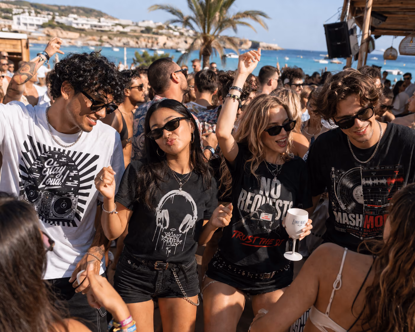 Group of young people dancing and enjoying a sunny beach party with ocean and palm tree in background.