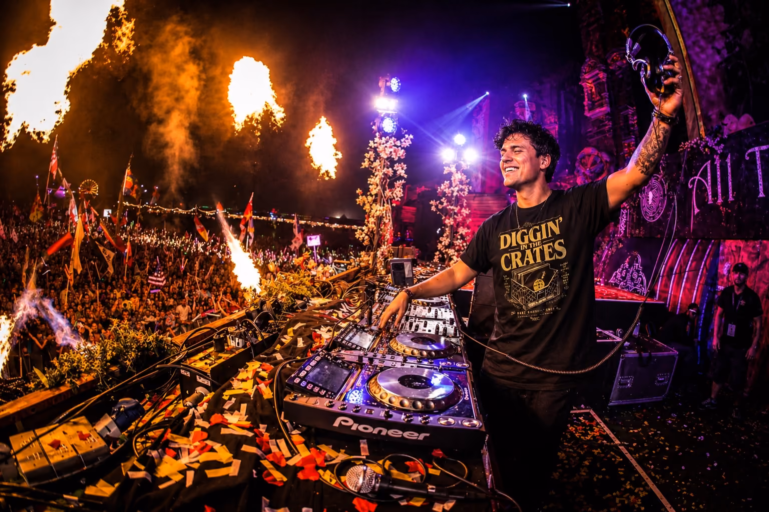 Smiling DJ playing music at a festival with crowd, flags, confetti, and flames in the background.