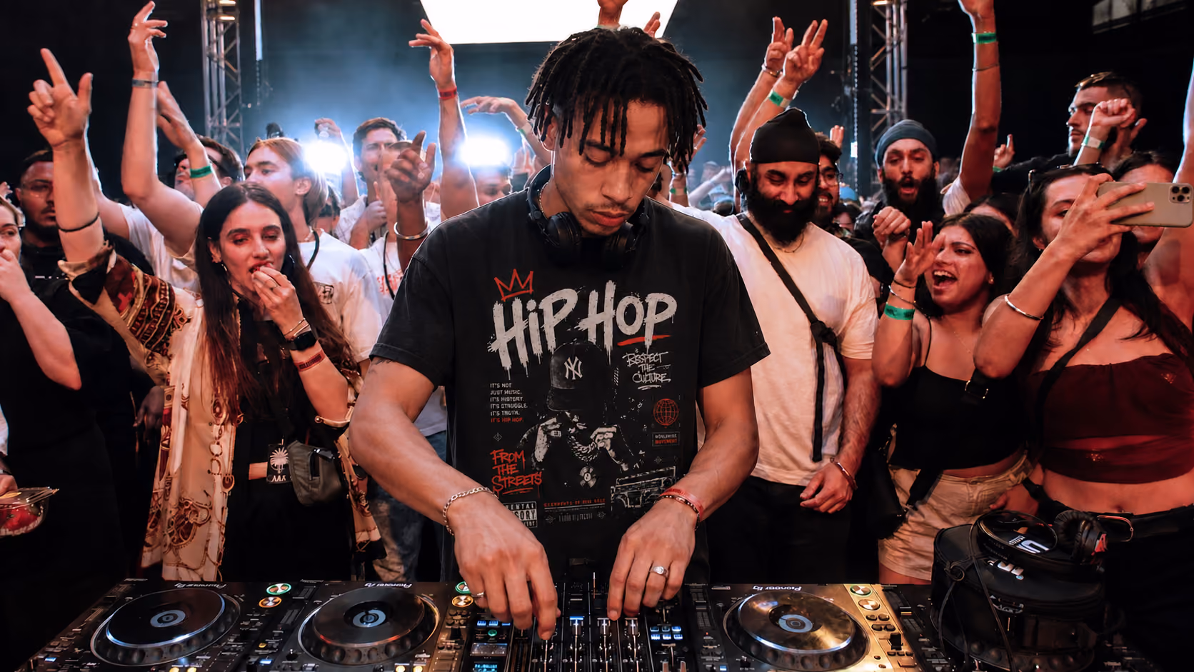 DJ wearing headphones and a hip hop shirt plays music on a mixer while a lively crowd cheers.