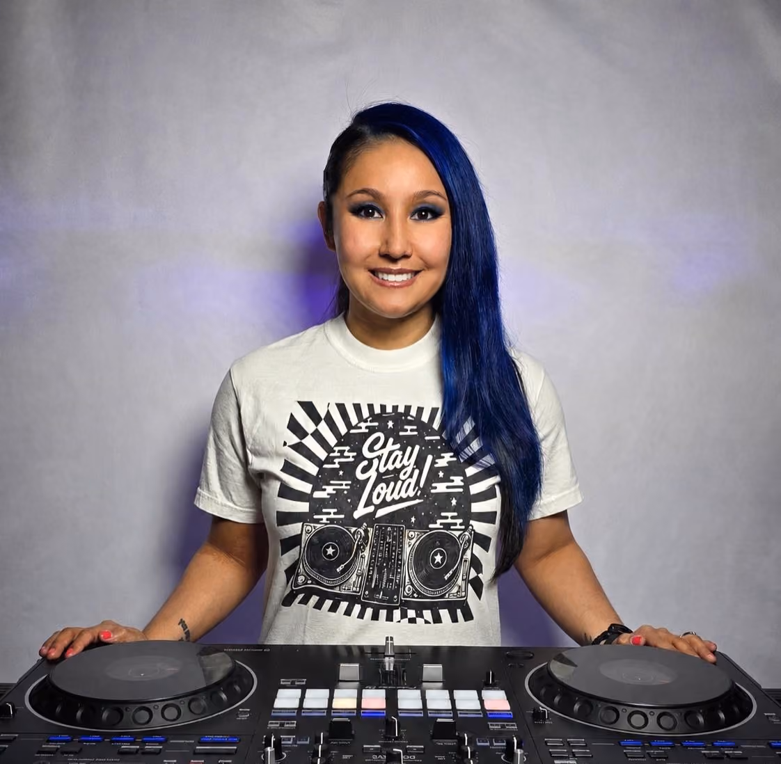 Smiling woman with long blue hair stands behind DJ controller wearing a Stay Loud! t-shirt.