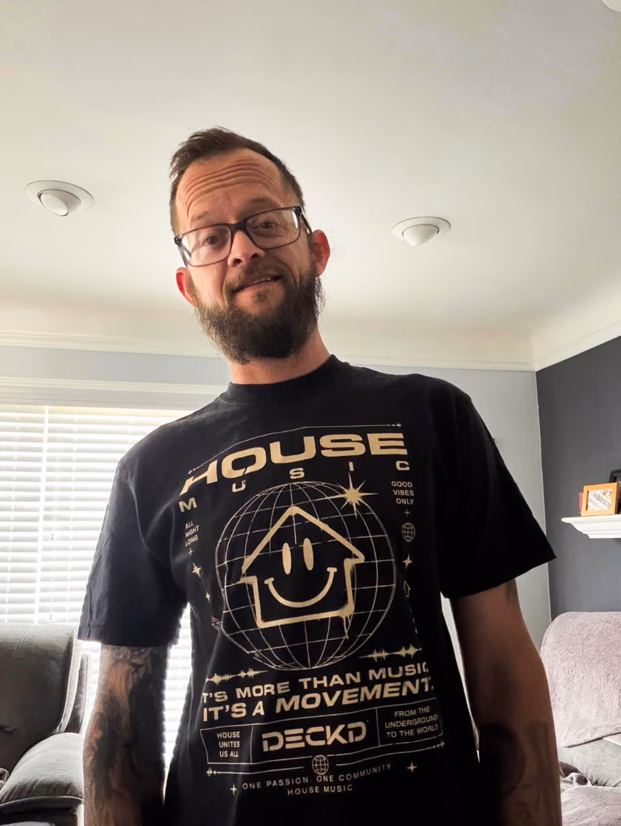 Man with glasses and beard wearing a black House Music t-shirt inside a living room.