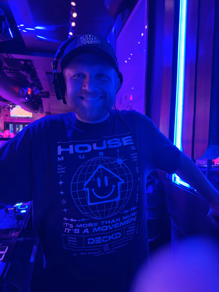 Smiling man wearing headphones and a house music t-shirt in a blue-lit room.