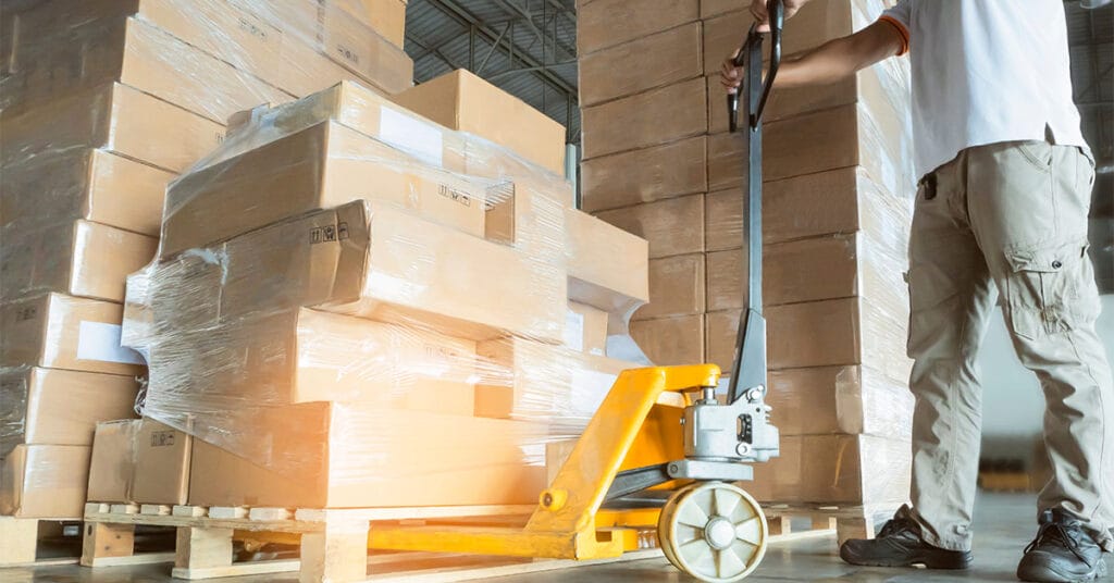 A person utilizing a pallet jack to move a large stack of cardboard boxes wrapped in plastic, likely prepared for LTL shipping. The boxes rest on a wooden pallet in a bustling warehouse setting. Wearing a white shirt and beige pants, the individual efficiently maneuvers the load.