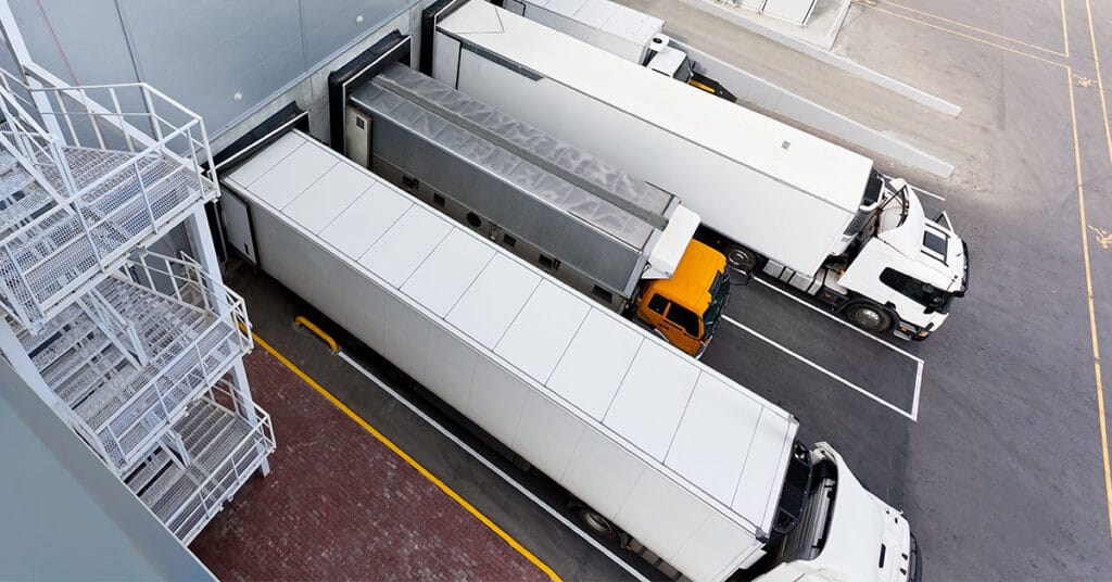 Aerial view of three large white semi-trucks involved in LTL shipping, backed into loading docks at a warehouse with a staircase on the left. One truck has a yellow cab, while the others have white cabs. The area is paved with some painted lines marking the space.