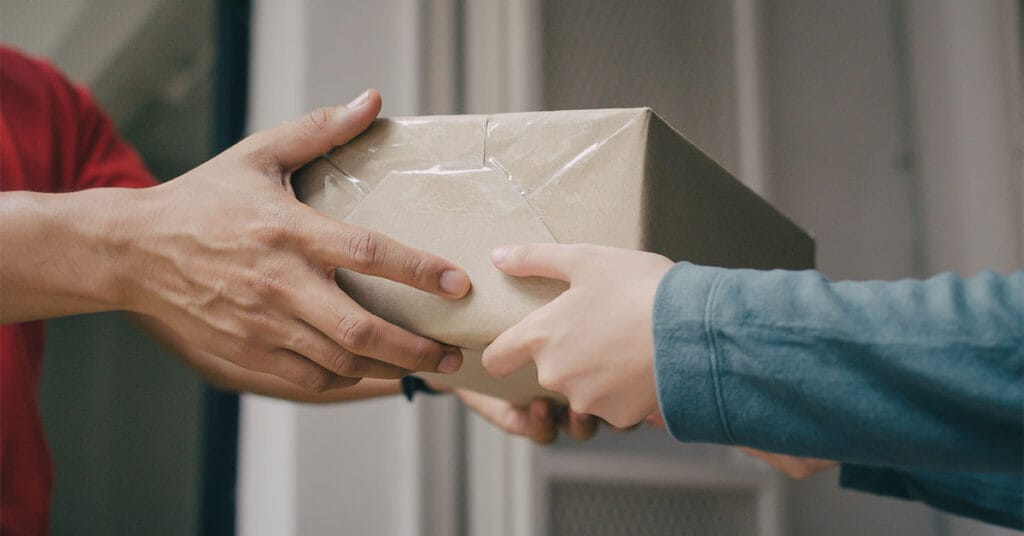 Close-up of two people exchanging a package, highlighting freight for ecommerce. One person wears a red shirt, handing a brown-wrapped parcel to another in a green jacket. The background is blurred, focusing attention on their hands and the package.