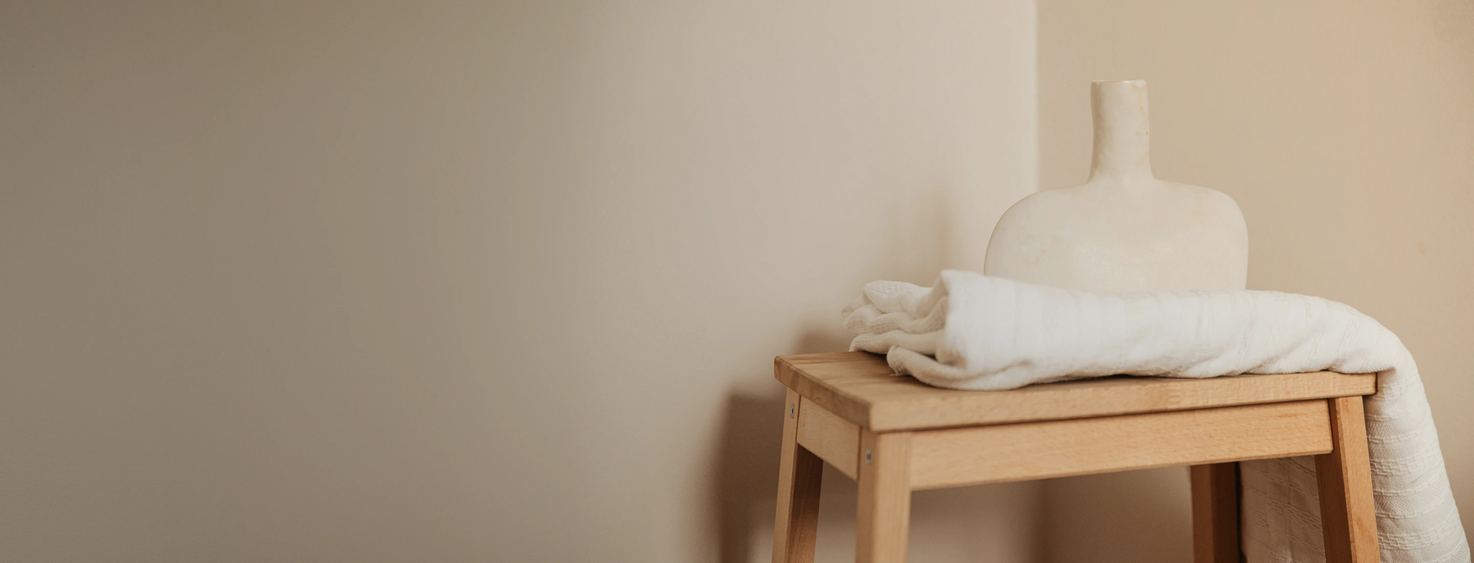 Folded white blanket and a beige ceramic vase on a wooden stool against a plain wall.