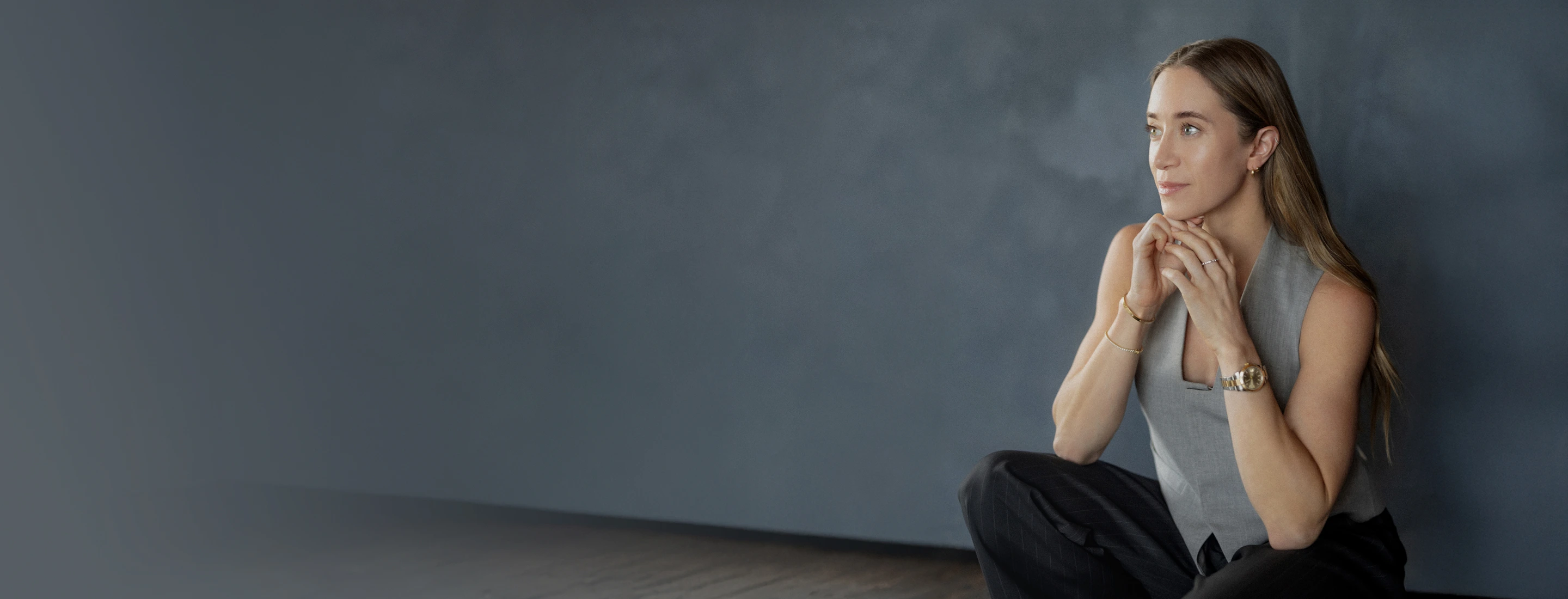 Kathryn Clorise sitting on floor against a gray wall, hands clasped near her face, looking right.