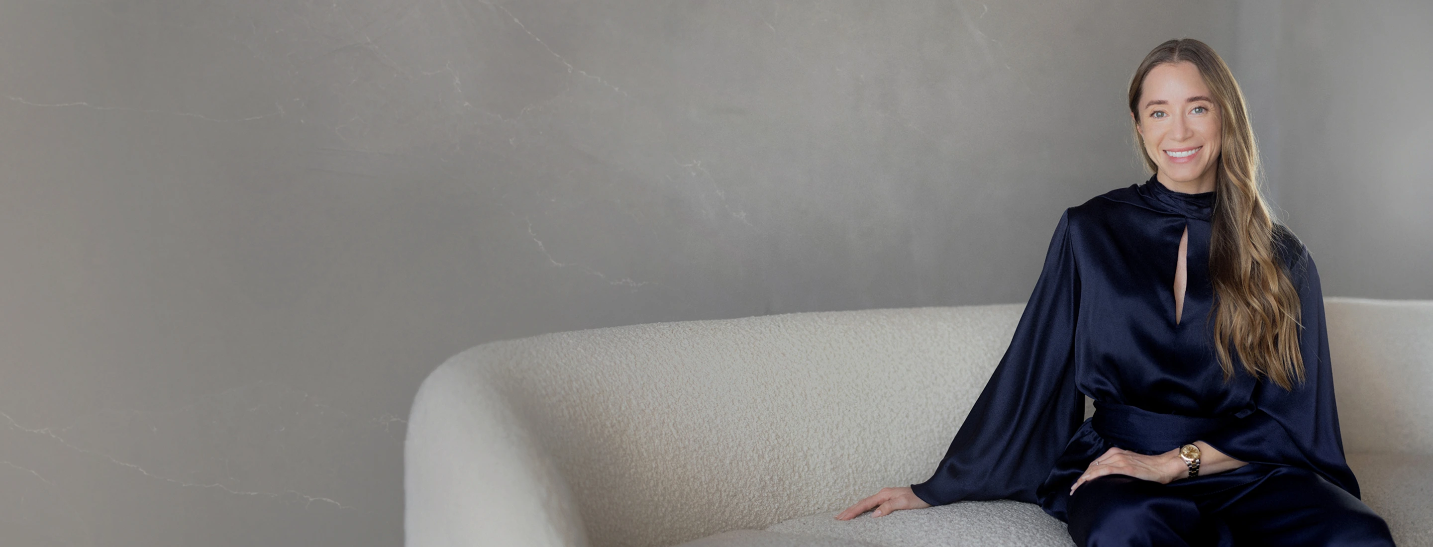 Kathryn Clorise in navy blue satin dress sitting on a cream sofa against a gray wall.