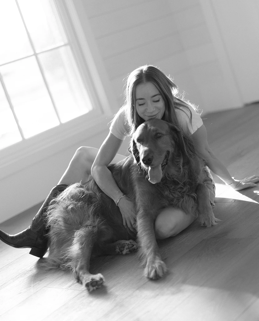 Kathryn Clorise sitting on floor hugging a large dog with tongue out near a window in bright room.