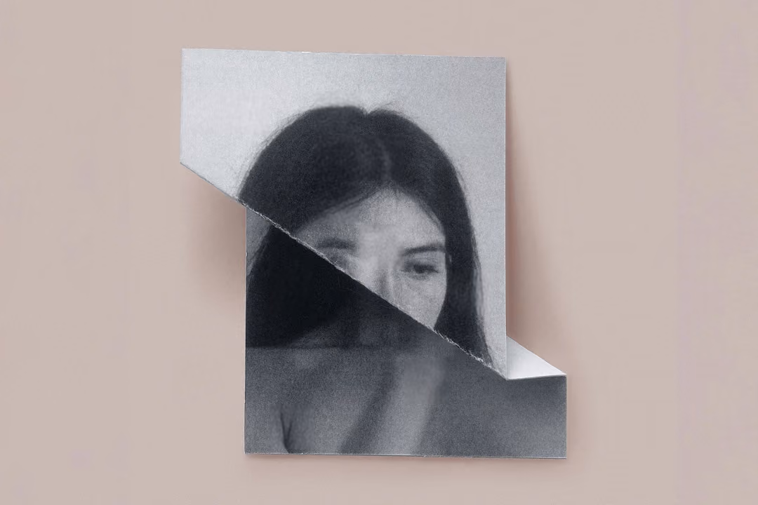 Black and white photo of a woman with long hair and bare shoulders partially obscured by torn paper.