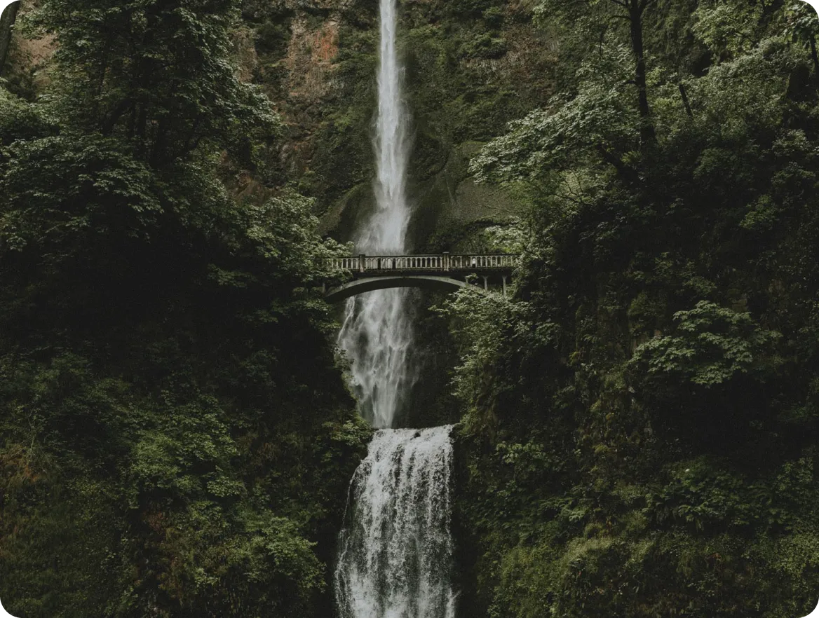 A tall waterfall cascading through a lush, forested canyon with a stone footbridge crossing partway down.