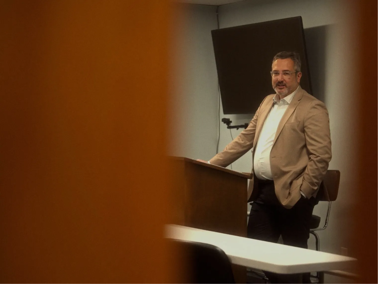 Tito, WRS's president, in a tan blazer, standing at a podium in a classroom, speaking to students with a video camera and acoustic panel visible in the background.