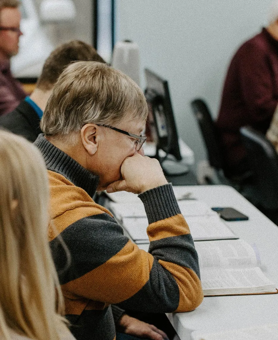 A student listening attentively during class, seated at a table with an open book, notes, and other classmates nearby.
