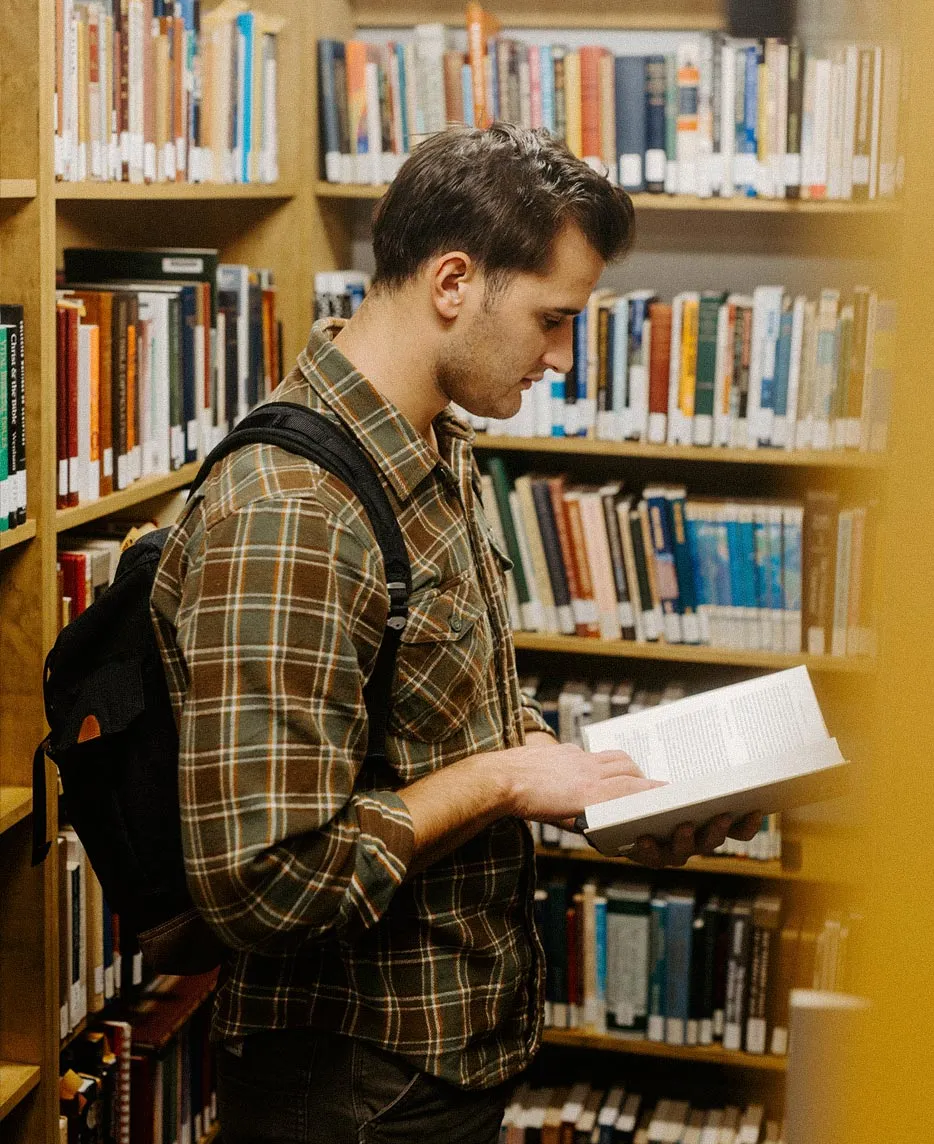 A student with a backpack reading a book while standing between library bookshelves.