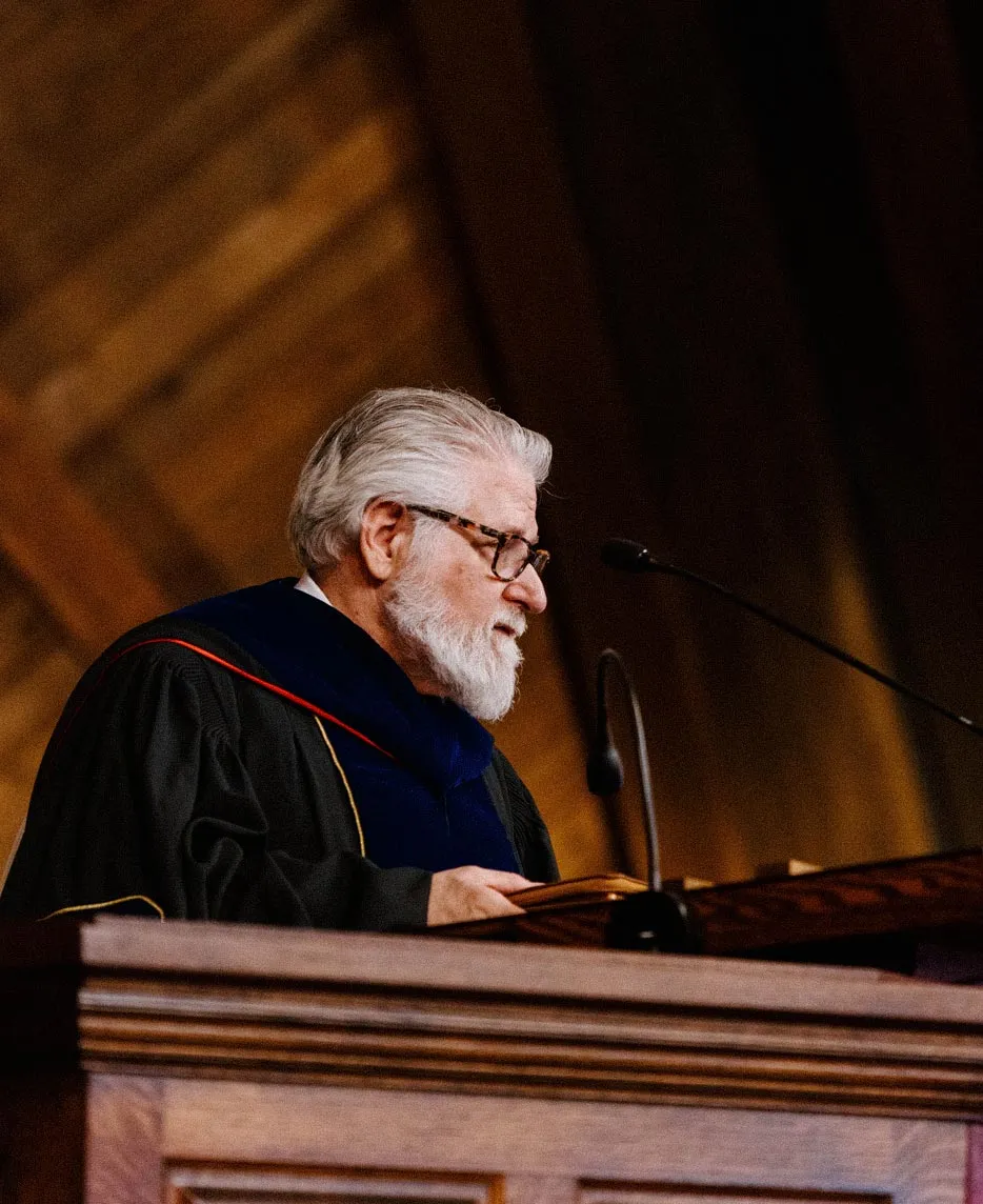 Kevin Backus in academic regalia and a blue hood speaking at a podium microphone during a seminary commencement ceremony.