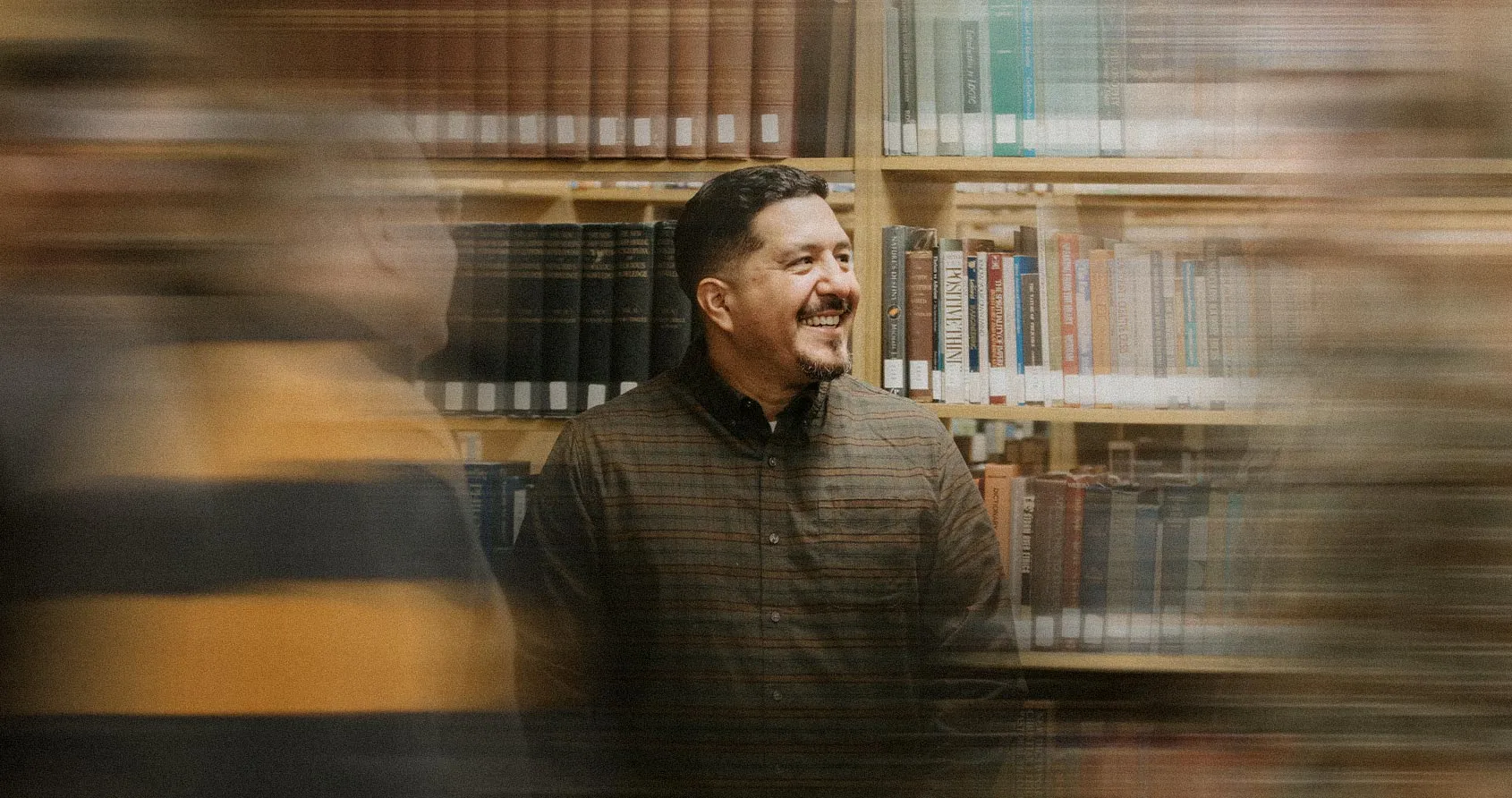 A man smiling while standing in a library among tall bookshelves, with motion blur suggesting a busy academic setting.