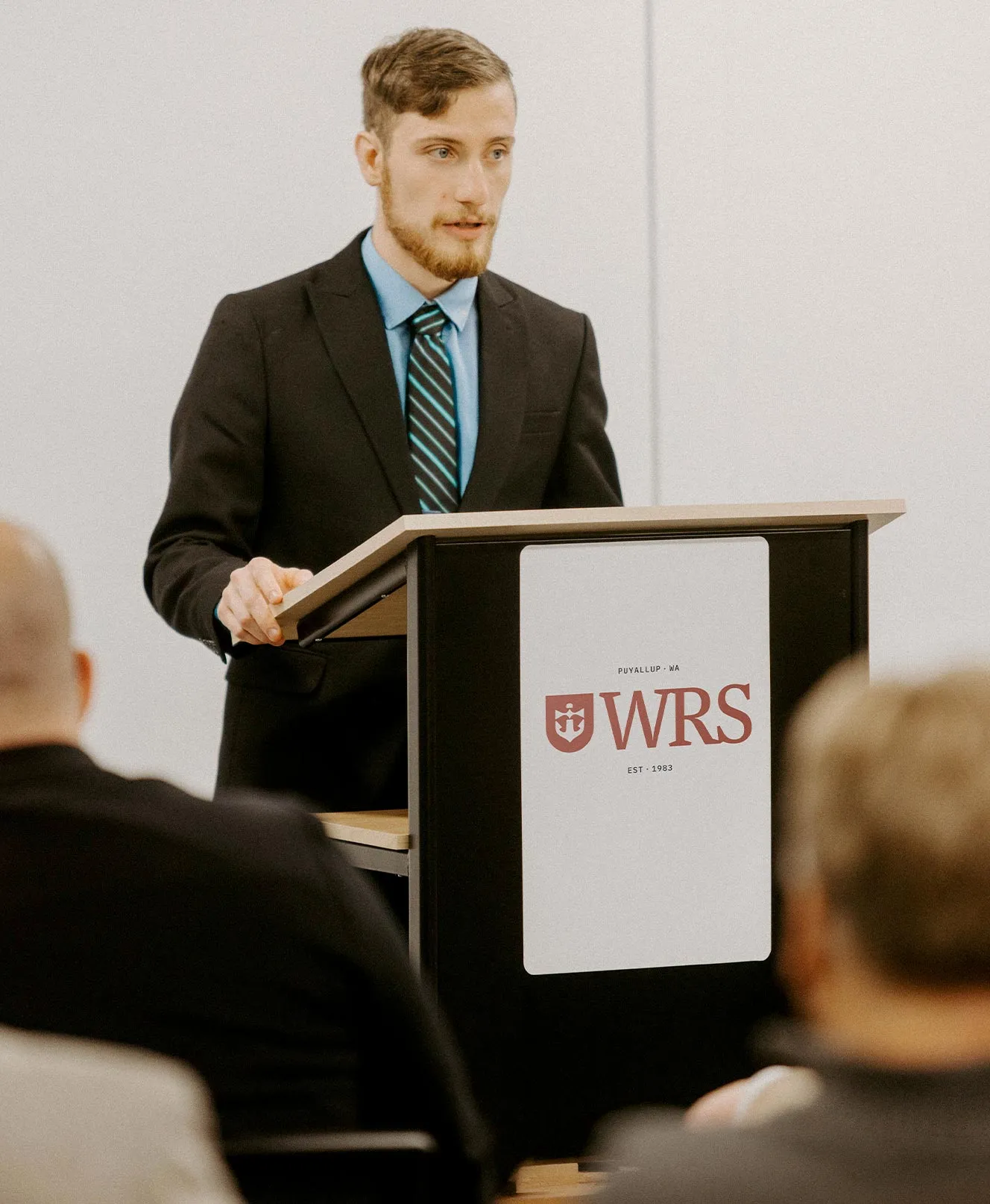 A man in a suit speaking at a podium branded with the Western Reformed Seminary crest, addressing an audience seated in the foreground.