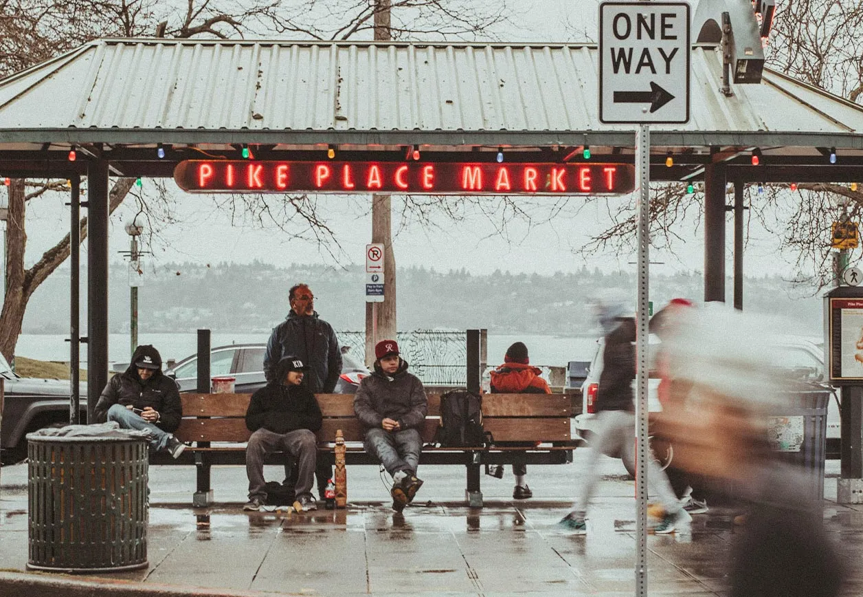 People sitting under the Pike Place Market sign in Seattle on a rainy day, with pedestrians passing by and Elliott Bay in the background.