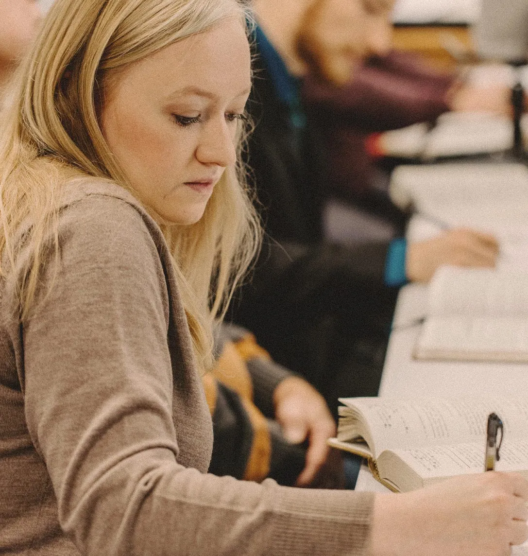 A student following along in an open book during a seminary classroom lecture, with other students seated nearby.