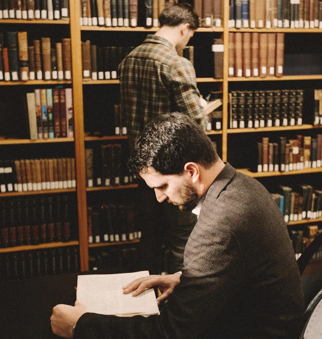 Two students reading in a seminary library, with one browsing tall wooden bookshelves lined with hardbound volumes in the background.