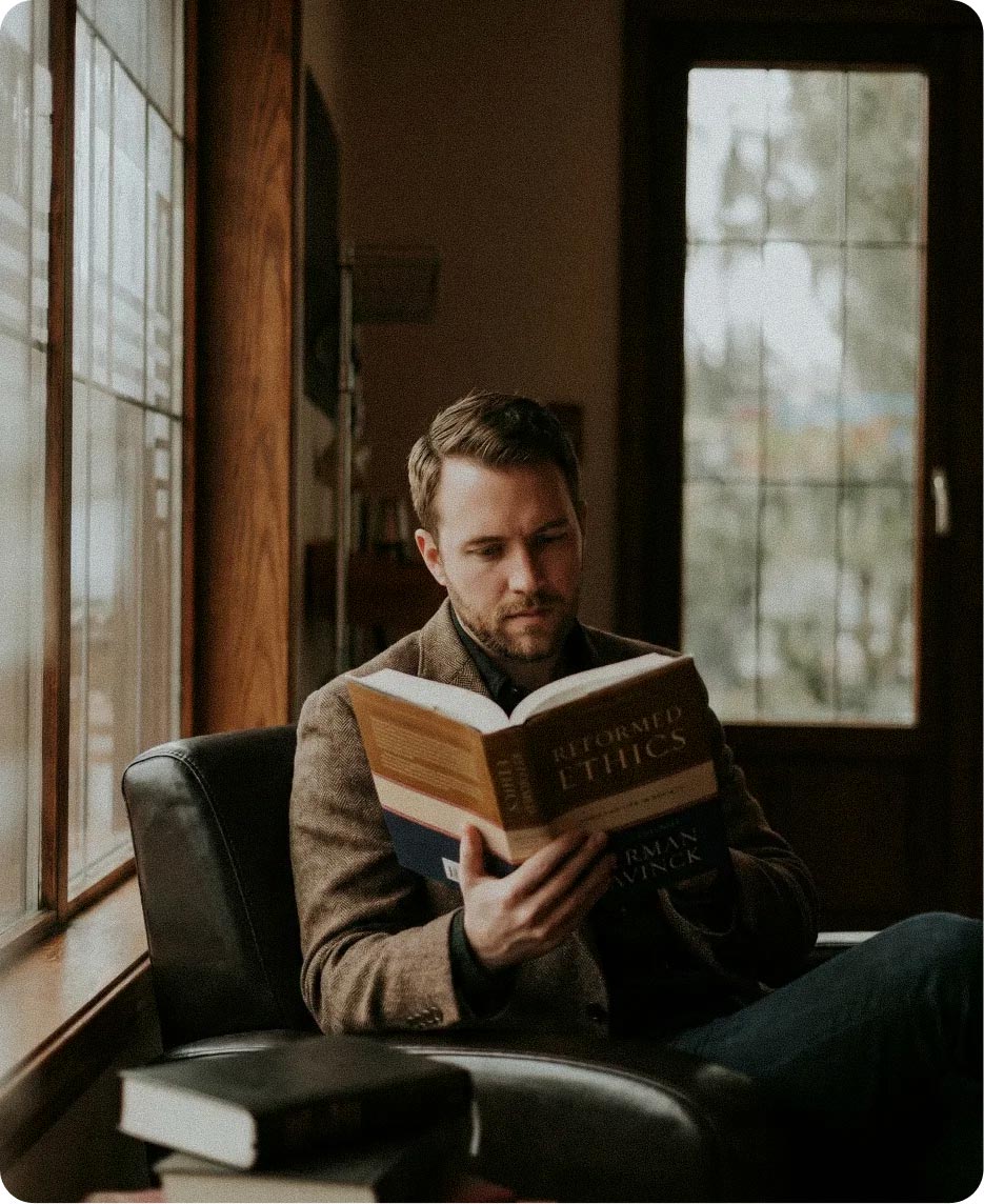 A man in a tweed blazer reading Herman Bavinck's Reformed Ethics while seated by a window in a study.