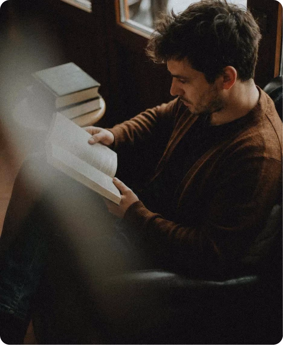 A man reading a book by a window, with a stack of books beside him in warm, softly lit surroundings.