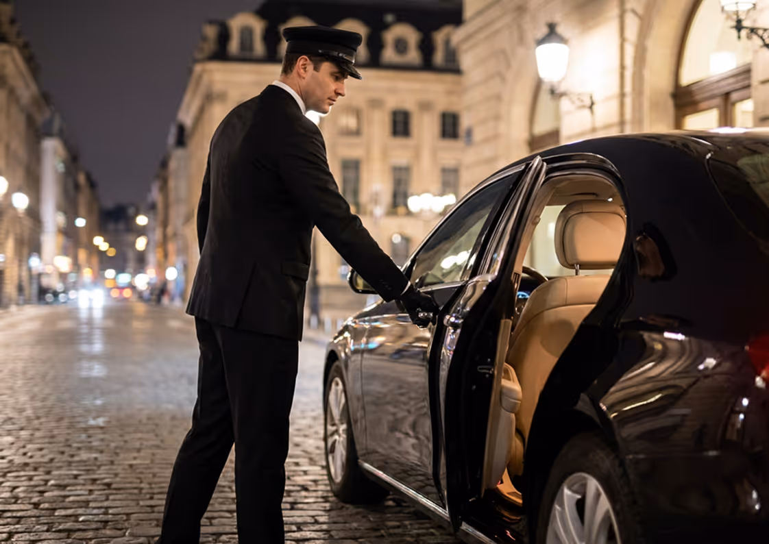 Chauffeur in uniform opening the door of a black luxury car on a cobblestone street at night.