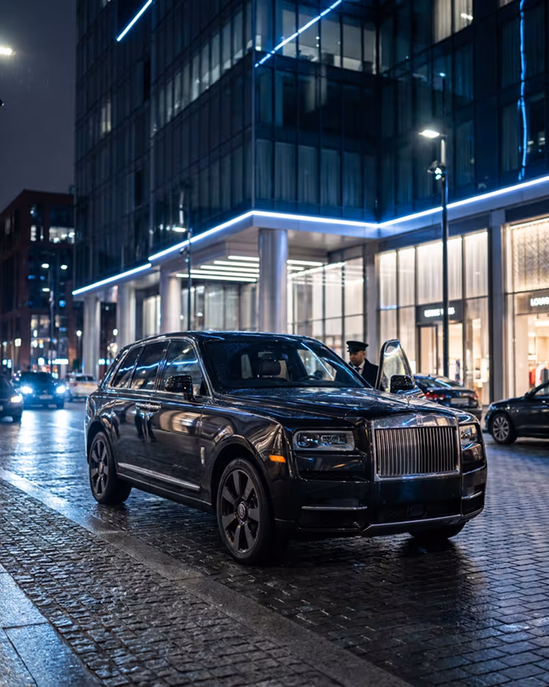 Black Rolls-Royce SUV parked on a wet city street at night with a driver opening the rear door.