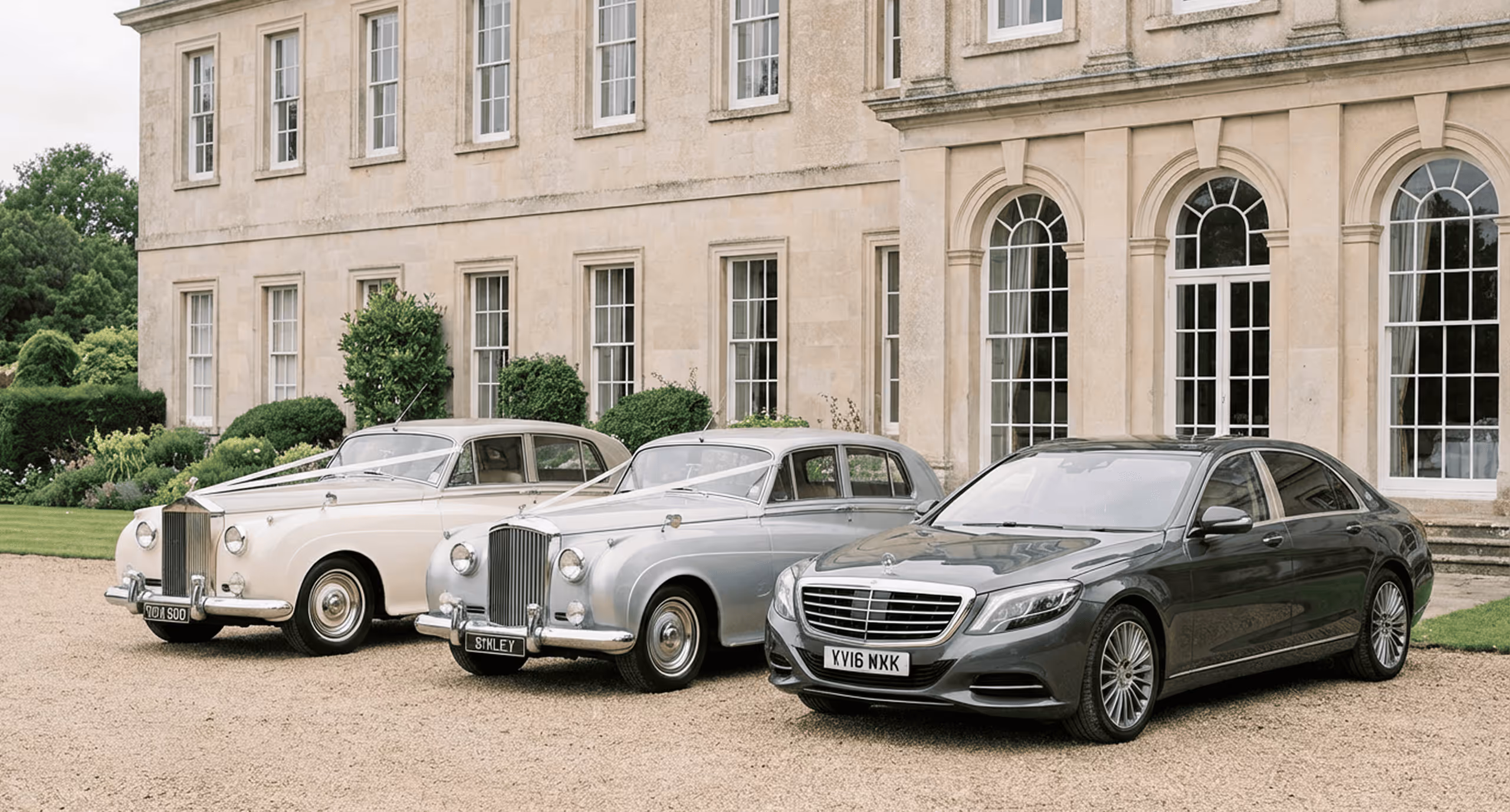 Two classic Rolls-Royce cars and one modern Mercedes-Benz sedan parked outside a large beige stone building.