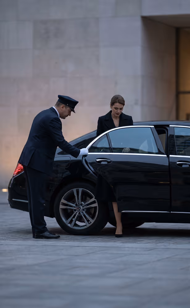 Chauffeur in uniform opening car door for woman exiting a black luxury vehicle on a city street.