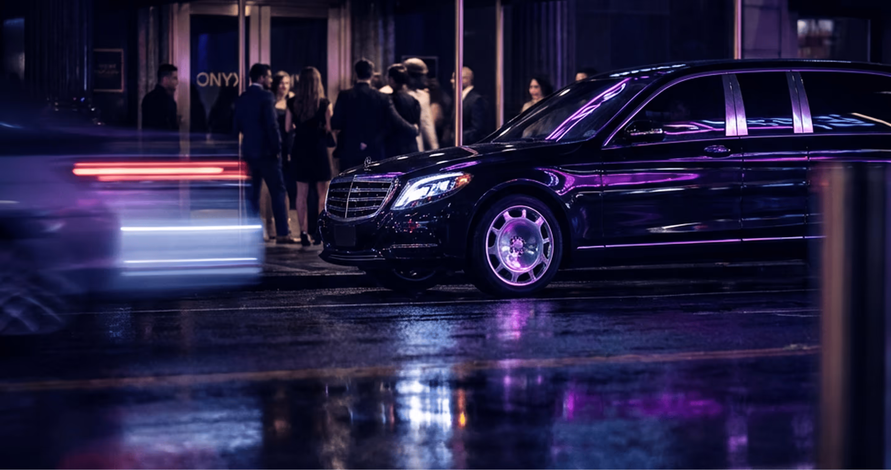 Black luxury Mercedes-Benz car parked on wet city street with blurred passing vehicle and people in background.