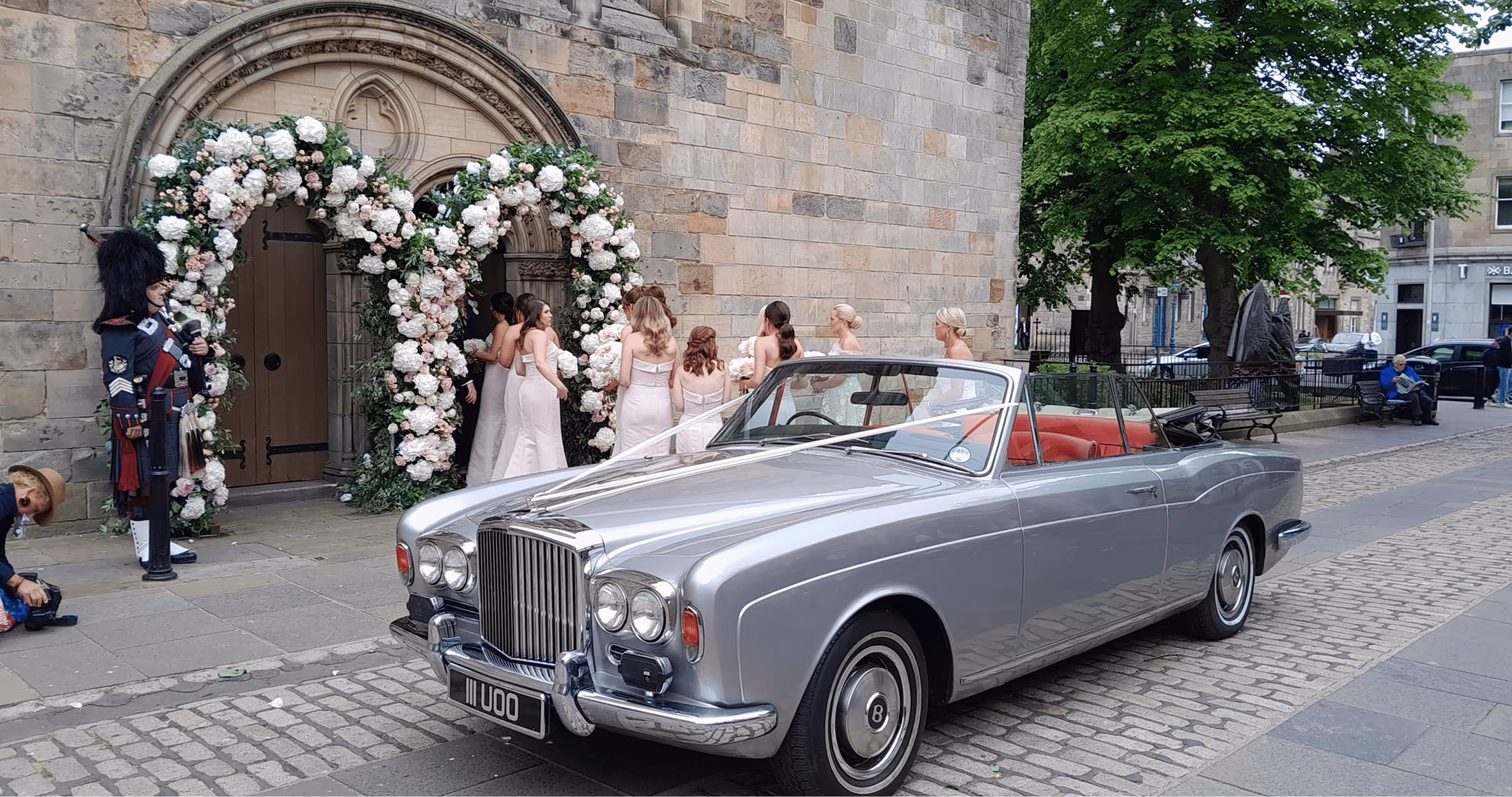 Silver vintage Bentley convertible parked outside a chapel with a floral wedding arch and bridesmaids in pink dresses
