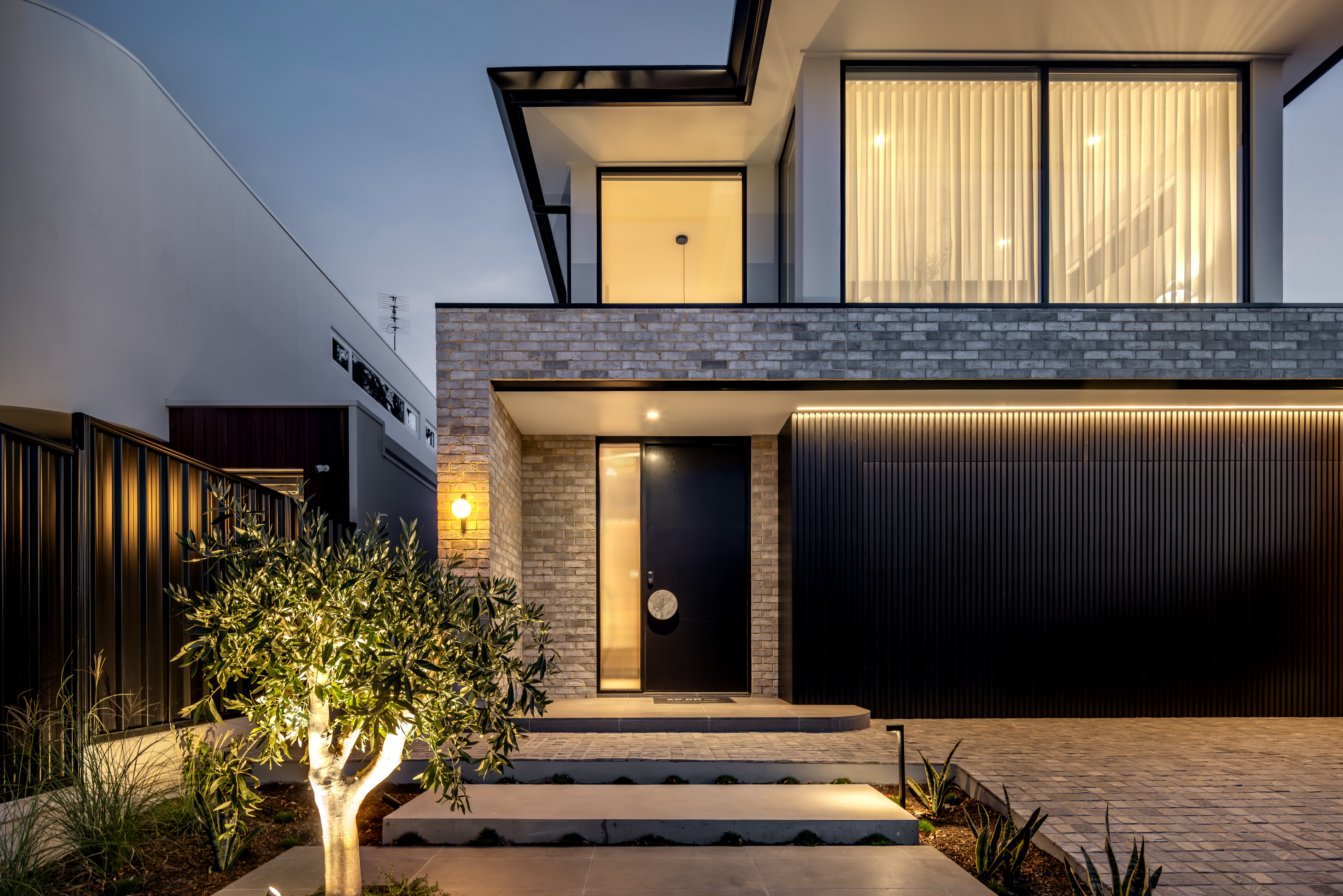 Street facade of 9 Netherby at dusk with grey brickwork, landscaped entry steps and an olive tree