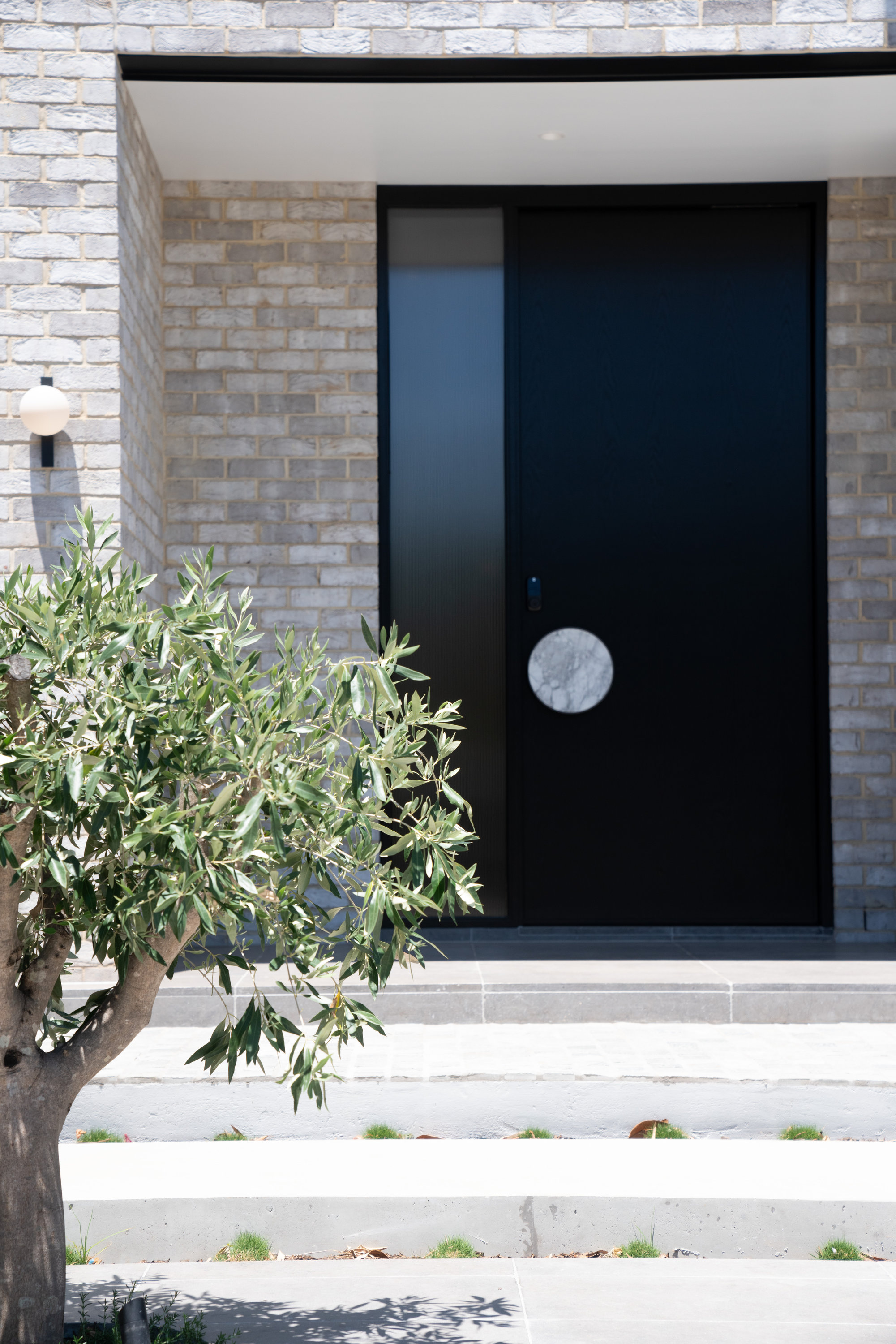 Black pivot entry door with a round marble handle plate, framed by brickwork and an olive tree at 9 Netherby
