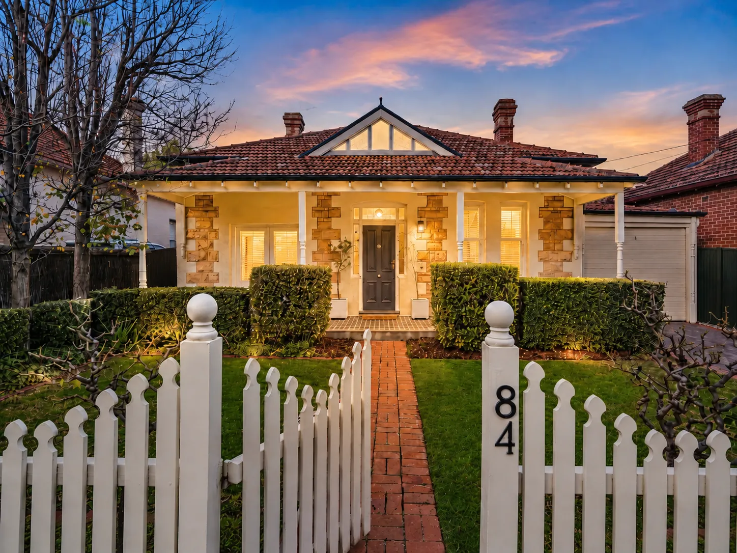 Symmetrical sandstone villa with a return verandah, picket fence and brick path at dusk at 84 Toorak Gardens