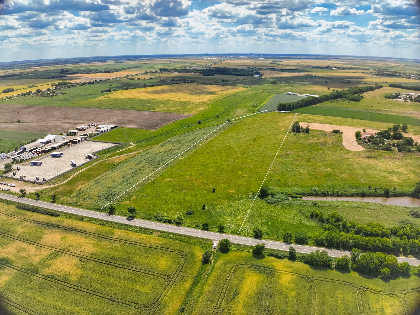 Aerial view of green fields, a winding river, farmland, road, and scattered buildings under blue sky.